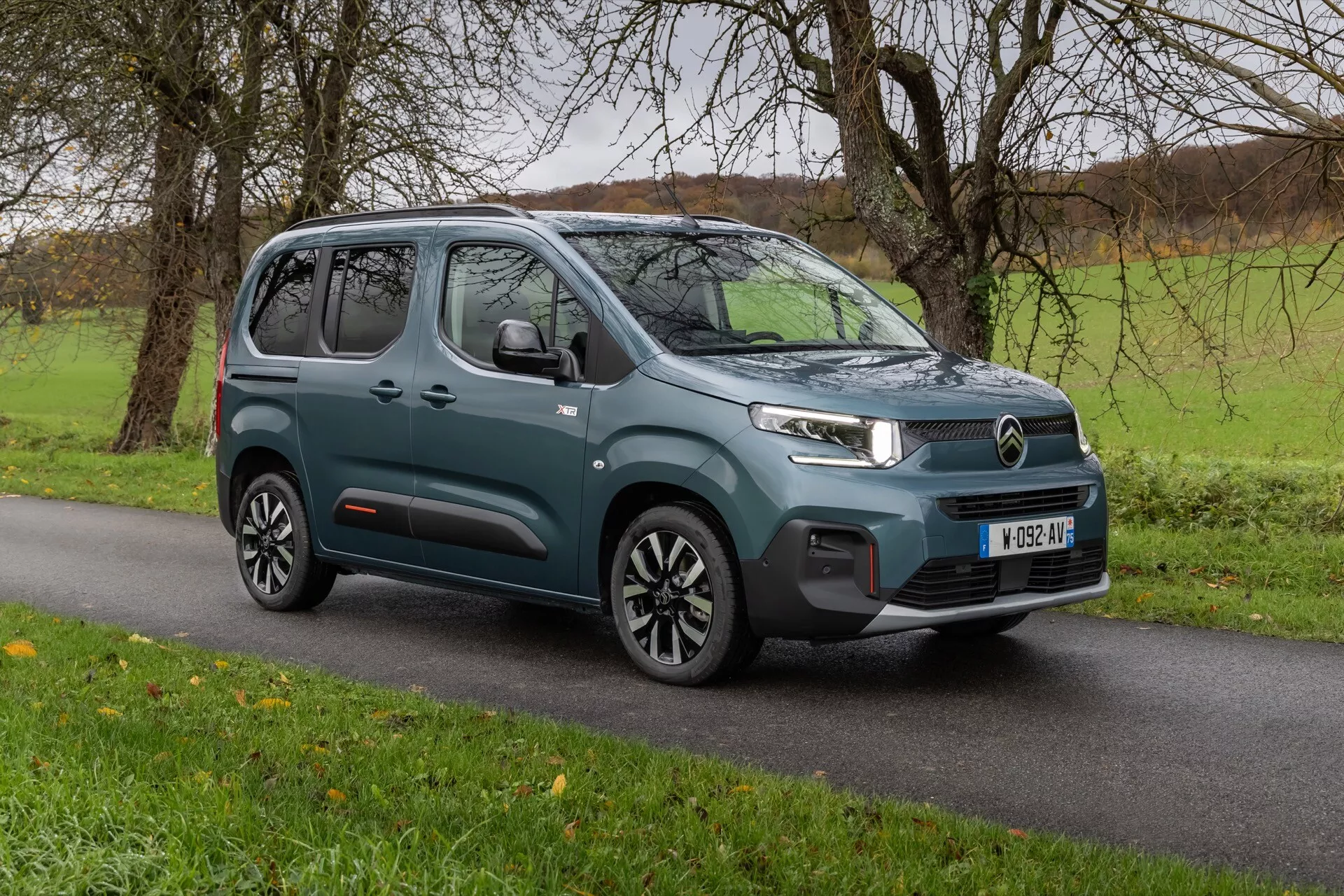 Facelift 3. generace Citroen Berlingo 2024, moderní kompaktní MPV na asfaltu s travnatým pozadím ukazujícím tříčtvrteční přední pohled, zdůrazňující LED světlomety, mřížku charakteristickou pro značku, nárazník s parkovacími senzory a elegantní boční konturové čáry s litými koly se šrouby ve dvoubarevném provedení.