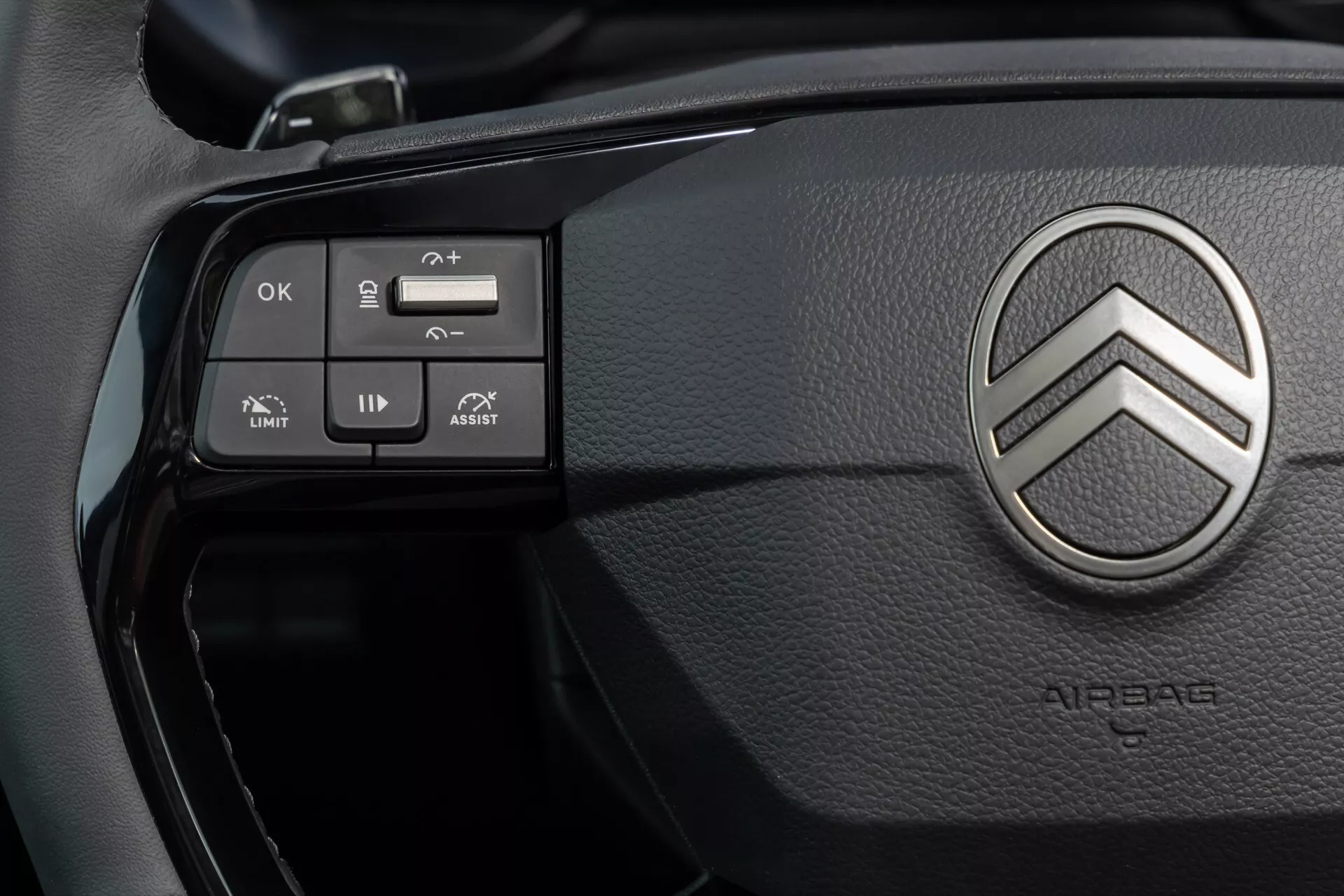 Detailní pohled na volant Citroënu Berlingo 2024 (facelift 3. generace) s kovovým dvojitým šípem, tlačítky pro ovládání tempomatu, audia, navigace v menu a asistenčními systémy jako omezovač rychlosti a asistent udržování jízdního pruhu