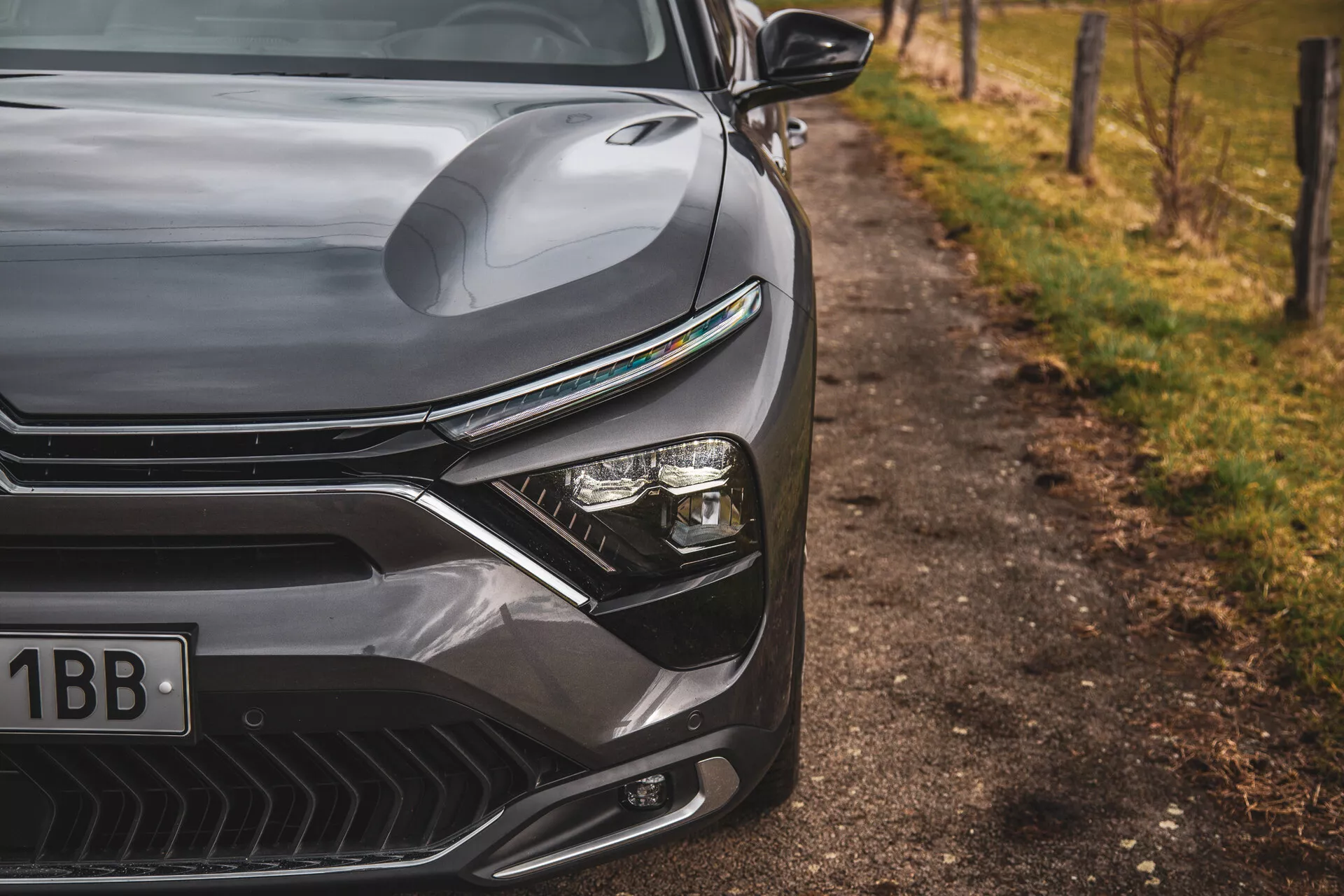 Detailní pohled na Citroen C5-X 2022 zobrazující elegantní design přední levé strany, včetně profilované kapoty, výrazné mřížky s horizontálními linkami, pokročilého systému LED světlometů a aerodynamicky tvarovaného předního nárazníku s otvory pro přívod vzduchu, proti rozmazanému přírodnímu pozadí u silnice.