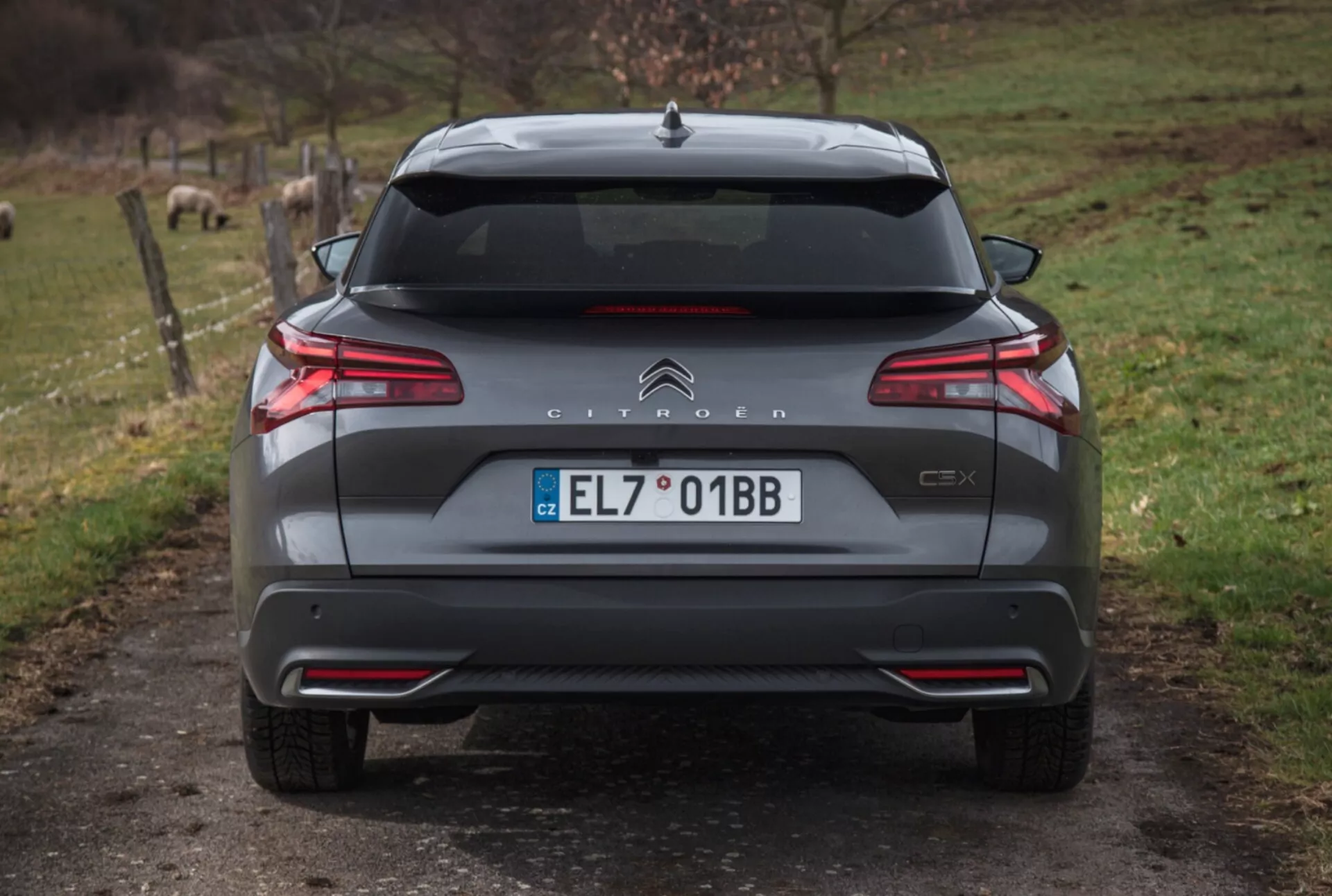Zadní pohled na Citroën C5X 2022 na štěrkové cestě s přírodním pozadím, předvádějící LED zadní světla, kontrolu vzdálenosti při parkování, dvojitý výfuk a další vnější detaily.