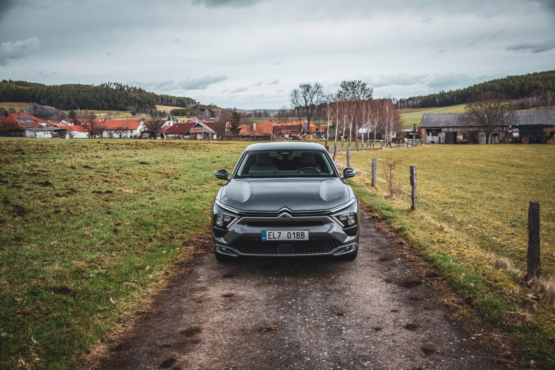 Čelní pohled na Citroën C5-X 2022 na úzké cestě v travnatém poli pod zataženou oblohou, předvádějící jeho elegantní design, složitou mřížku s emblémem, úhlové LED světlomety, sochařské linie kapoty a dodatečné přední osvětlovací prvky, registrovaný v České republice.