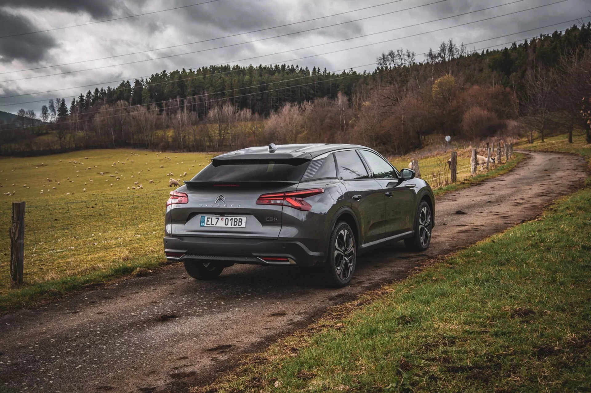Moderní hatchback Citroen C5-X 2022 zaparkovaný na nezpevněné venkovské silnici, pohled zezadu a ze strany ukazuje aerodynamický design s křídlem a žraločí ploutví na střeše, štíhlá vodorovná zadní světla a velká kola s mnoha paprsky, což naznačuje výkon a luxus v mimo-městském prostředí.