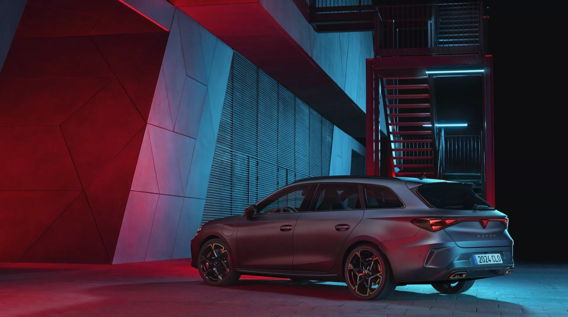 Cupra Leon 2024 (1. generace facelift) fotografovaný ze zadního, tříčtvrtečního úhlu zdůrazňujícího stranu pro pasažéry. Dramatické noční osvětlení zdůrazňuje designové prvky vozu, včetně sportovních zadních světlometů, zadního nárazníku, agresivního střešního spoileru a výkonově orientovaných ráfků na pozadí městského průmyslového prostředí.