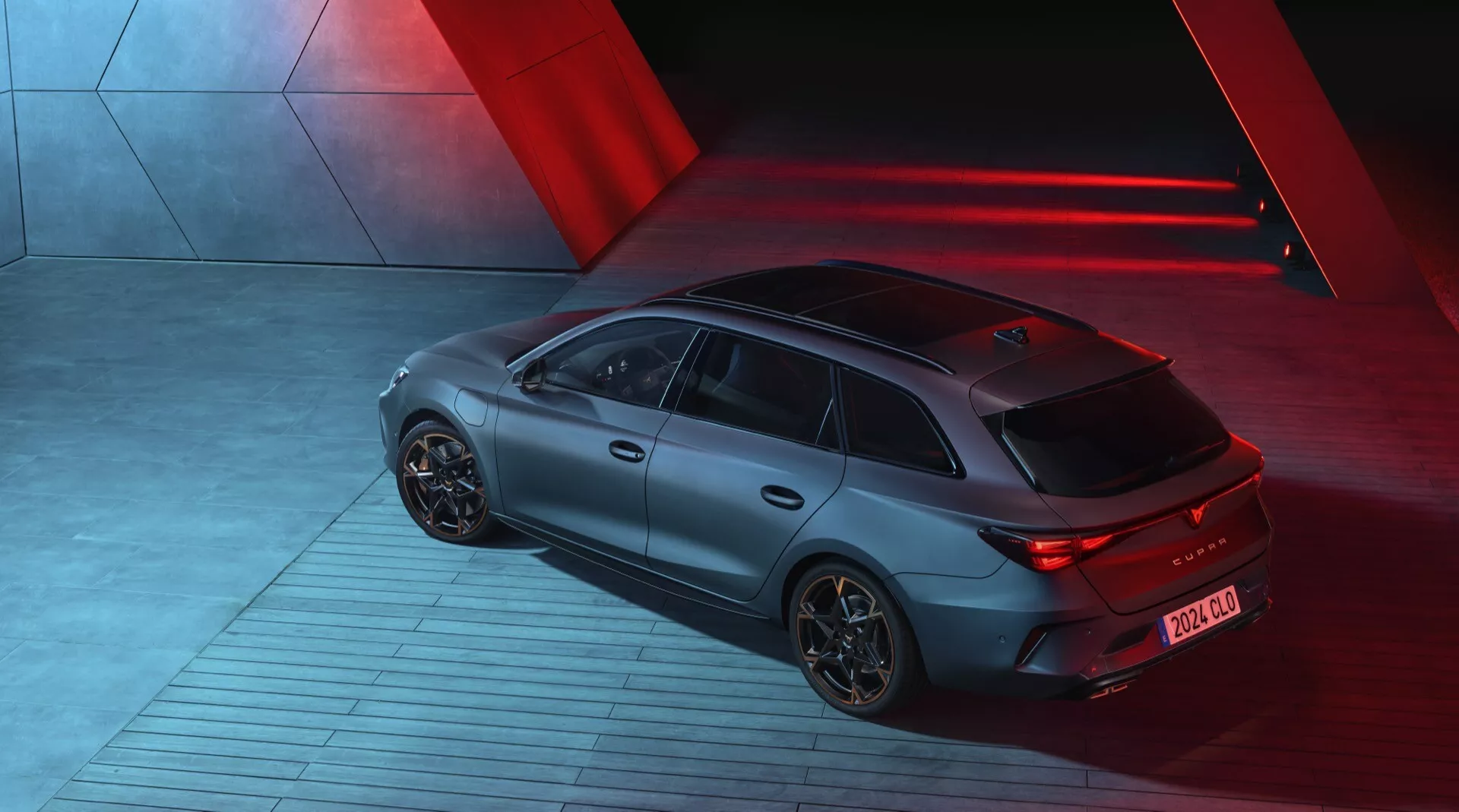 Pohled z výšky na model Cupra Leon 2024 (1. generace facelift) pořízený v noci, zobrazující zadní a pravou stranu sportbacku. Auto je osvětleno dramatickými červenými směrovými světly, která vytváří elegantní, futuristickou atmosféru. Má matný povrch, osvětlená LED zadní světla a aerodynamická slitinová kola. Vozidlo stojí na vzorovaném průmyslovém povrchu, který naznačuje pohyb. Viditelné technologické prvky zahrnují zadní difuzor a krátkou anténu směrem dozadu pro komunikační technologie.