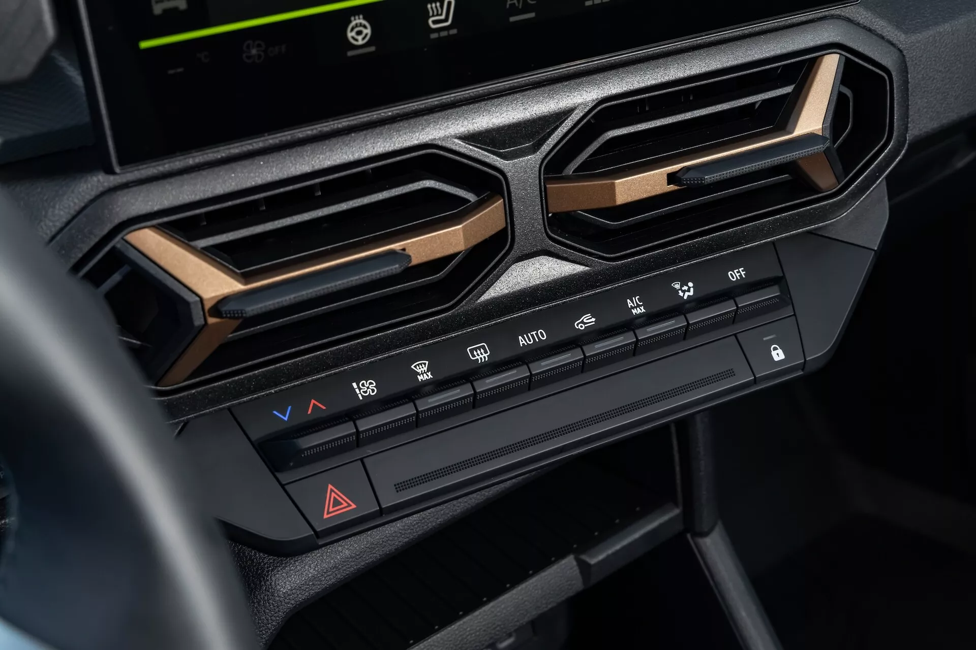 Detailní pohled na systém klimatizace v Dacia Duster 3. generace 2024 na středovém panelu pod obrazovkou infotainmentu, zahrnující ovládání teploty a rychlosti ventilátoru, funkce rozdělení vzduchu, odtávání, tlačítko výstražných světel a možnosti klimatizace, obklopené úhlovými ventilačními mřížkami a zdůrazněné metalickým lemem pro moderní estetiku interiéru