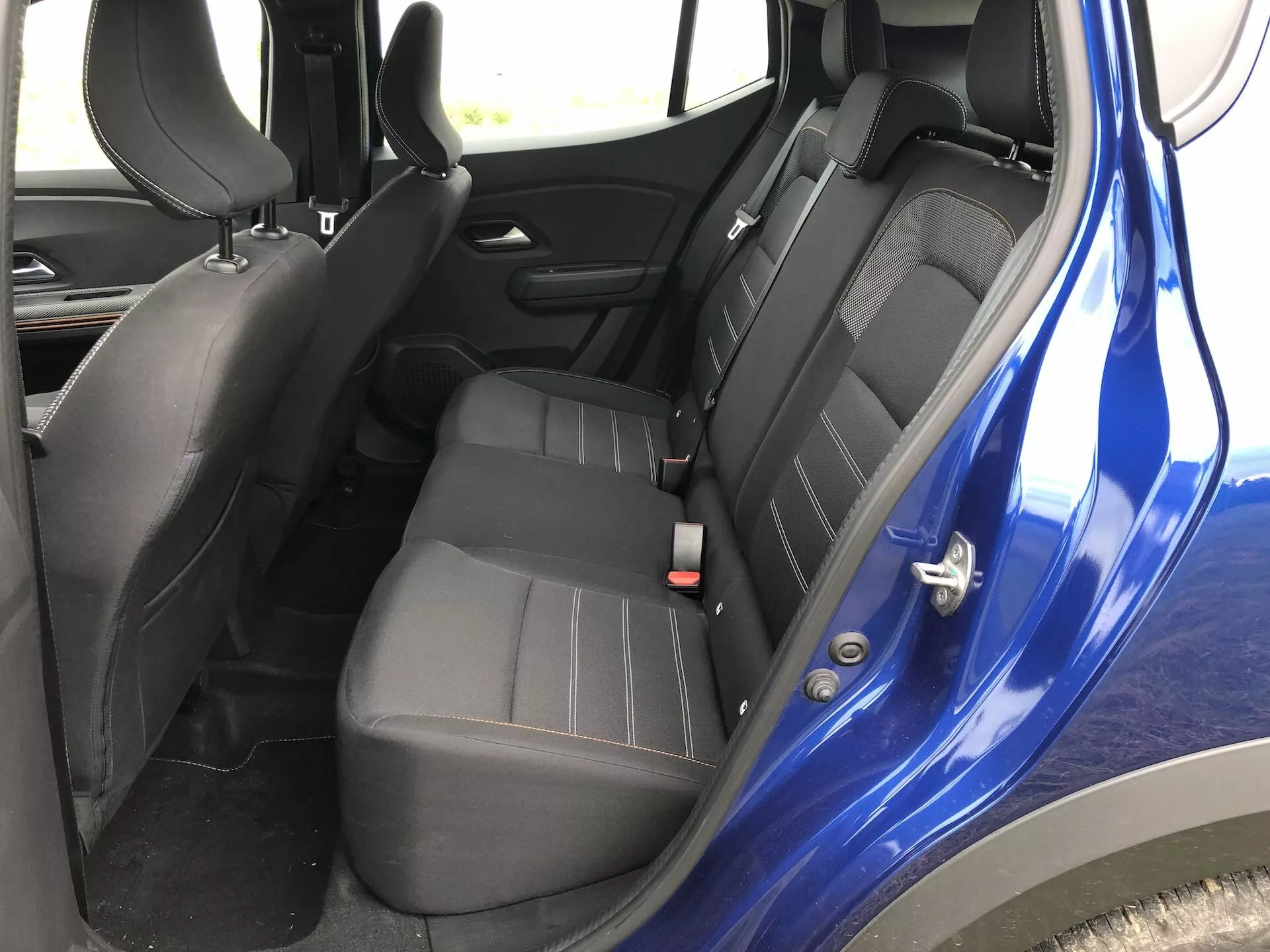 Interiér Dacia Sandero-2020 (Sandero-Stepway) se zaměřením na prostor zadních sedadel. Pohled z mírně vyšší úrovně sedadel, ukazující pravou stranu s bezpečnostními pásy, klikou dveří a tmavou barevnou kombinací, obsahující černý plast a tmavě šedé nebo černé čalounění. Zdůrazňuje konfiguraci sedadel, bezpečnostní prvky, jako jsou nastavitelné bezpečnostní pásy a možná ISOFIX úchyty, ovládání elektrického okna na panelu dveří a celkový design interiéru.