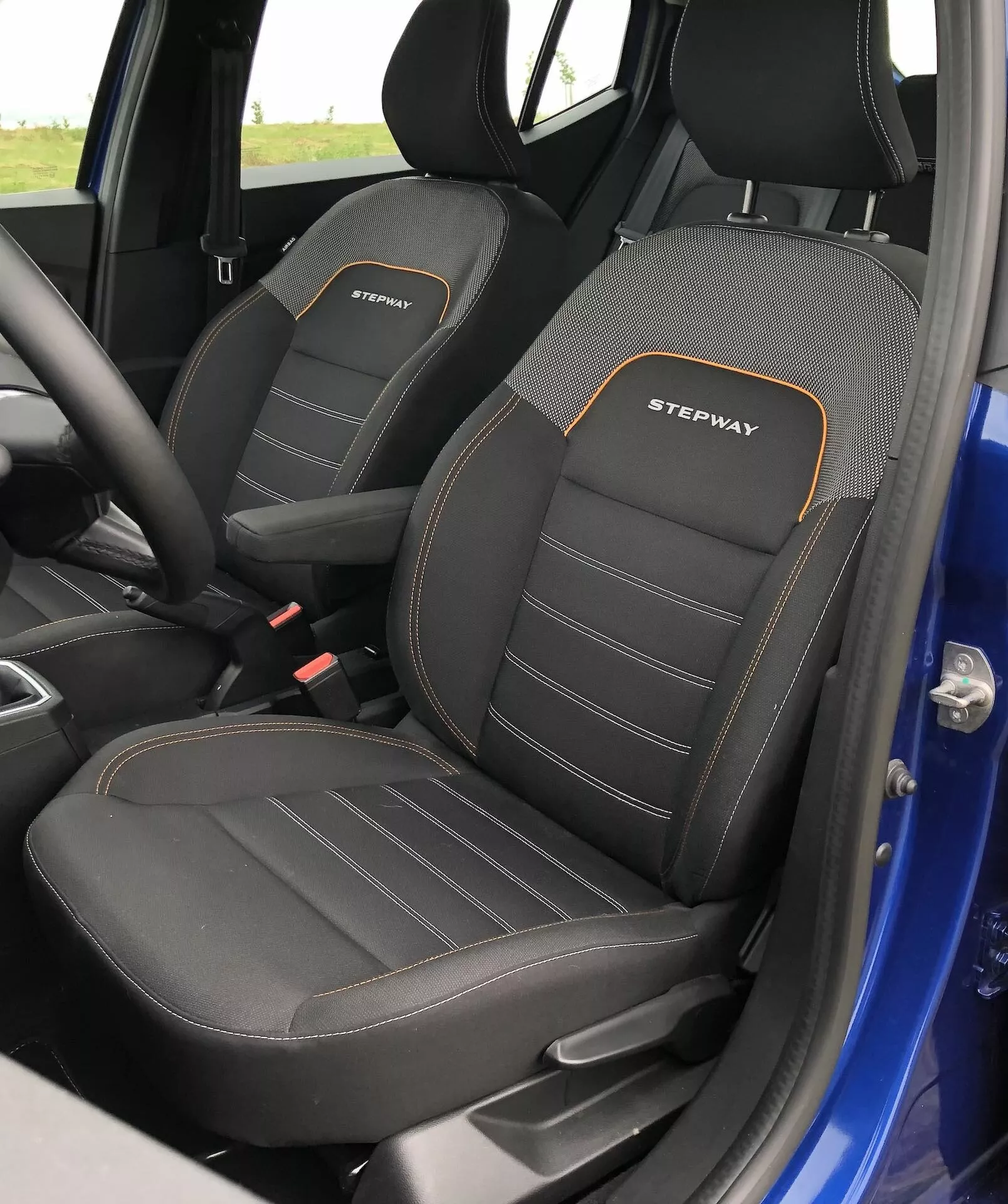 Detailní pohled na interiér předních sedadel Dacia Sandero 2020 Stepway, kde je vidět řidičovo sedadlo s výraznými bočními polštářky a výšivkou 'STEPWAY' v bílé barvě s oranžovým obrysem. Sedadla mají dvoubarevný design, převážně černý s texturovaným vzorem a viditelným dvojitým šitím. Fotografie zachycuje boční profily sedadel z mírně zvýšeného úhlu, což odhaluje horní část a část sedací plochy, což naznačuje sportovní a odolný výbavu. Za sedadly je viditelná malá část modrého vnějšího rámu dveří.
