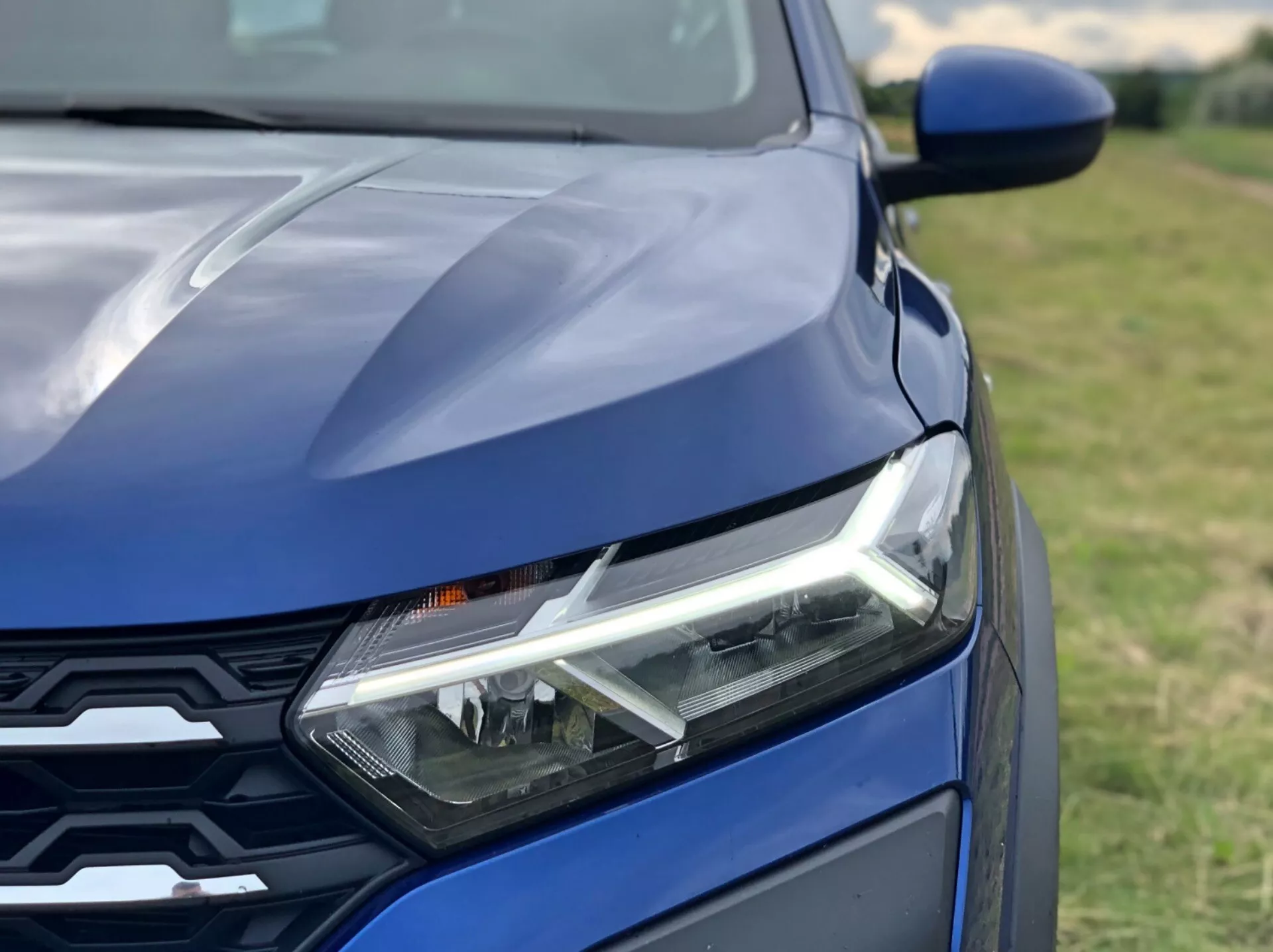 Blízký záběr přední části tmavě modrého vozu Dacia Sandero 2020 (Sandero Stepway) se zřetelným levým světlometem s denními LED světly, částí kapoty se sportovními liniemi a moderní přední maskou s geometrickým vzorem. Fotografováno ze šikmého úhlu nad úrovní světel s malou hloubkou ostrosti, která zdůrazňuje komponenty vozu.
