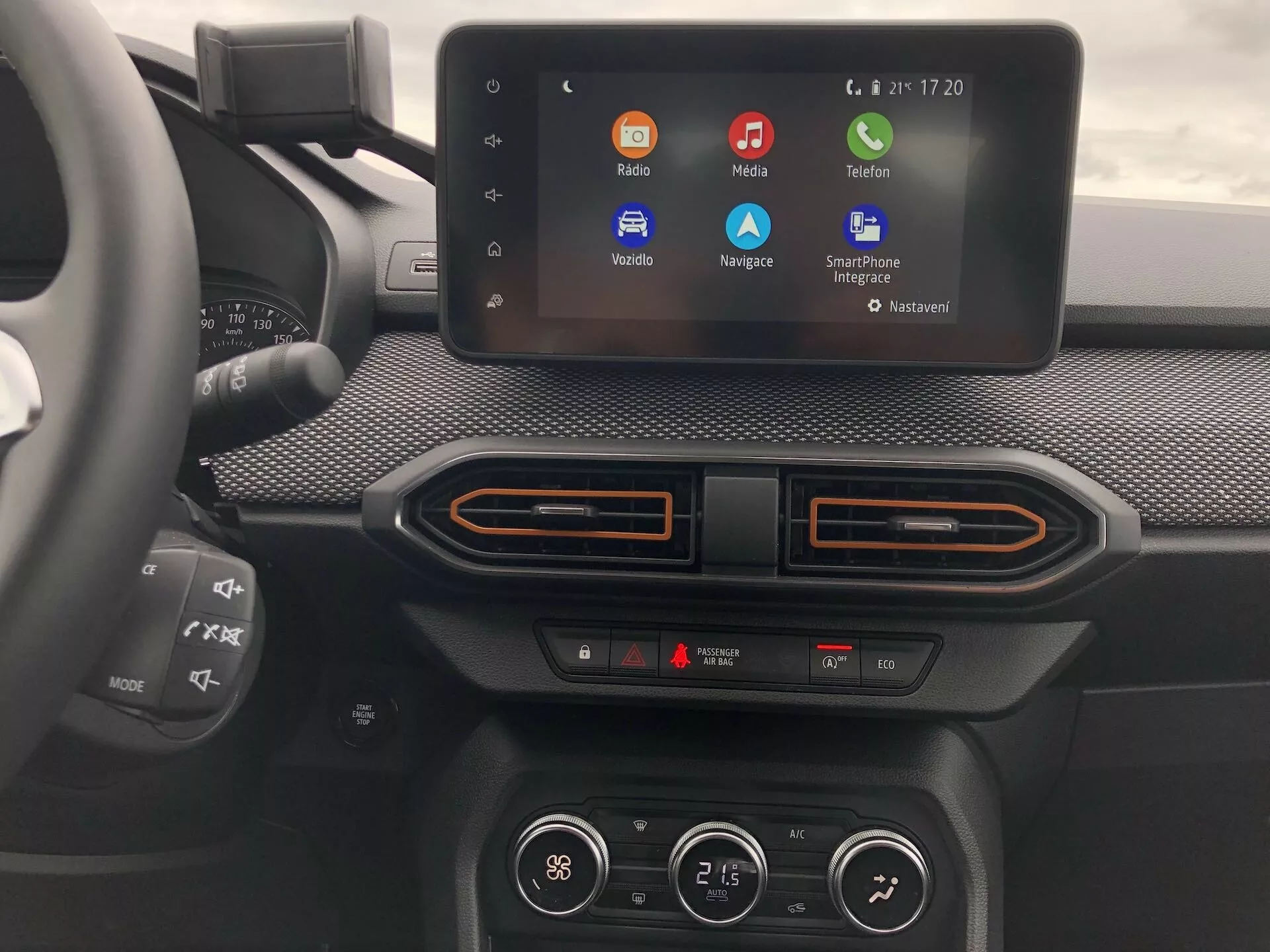 Detailní záběr na středovou konzoli Dacia Sandero Stepway 2020, zobrazující moderní dotykový infotainment systém s barevnými ikonami aplikací, umístěný v úrovni očí z pohledu pasažéra. Displej infotainmentu, zasazený do texturované palubní desky se zaoblenými rohy, naznačuje integraci smartphonu prostřednictvím Apple CarPlay a Android Auto. Níže jsou vidět dvouzónové klima ovladače se dvěma ukazateli teploty a unikátními horizontálními ventilačními otvory s barevnými akcenty. Částečný pohled na multifunkční volant vlevo. Displej ukazuje pokročilé technologické funkce jako hlasové příkazy, navigaci a streamování médií.