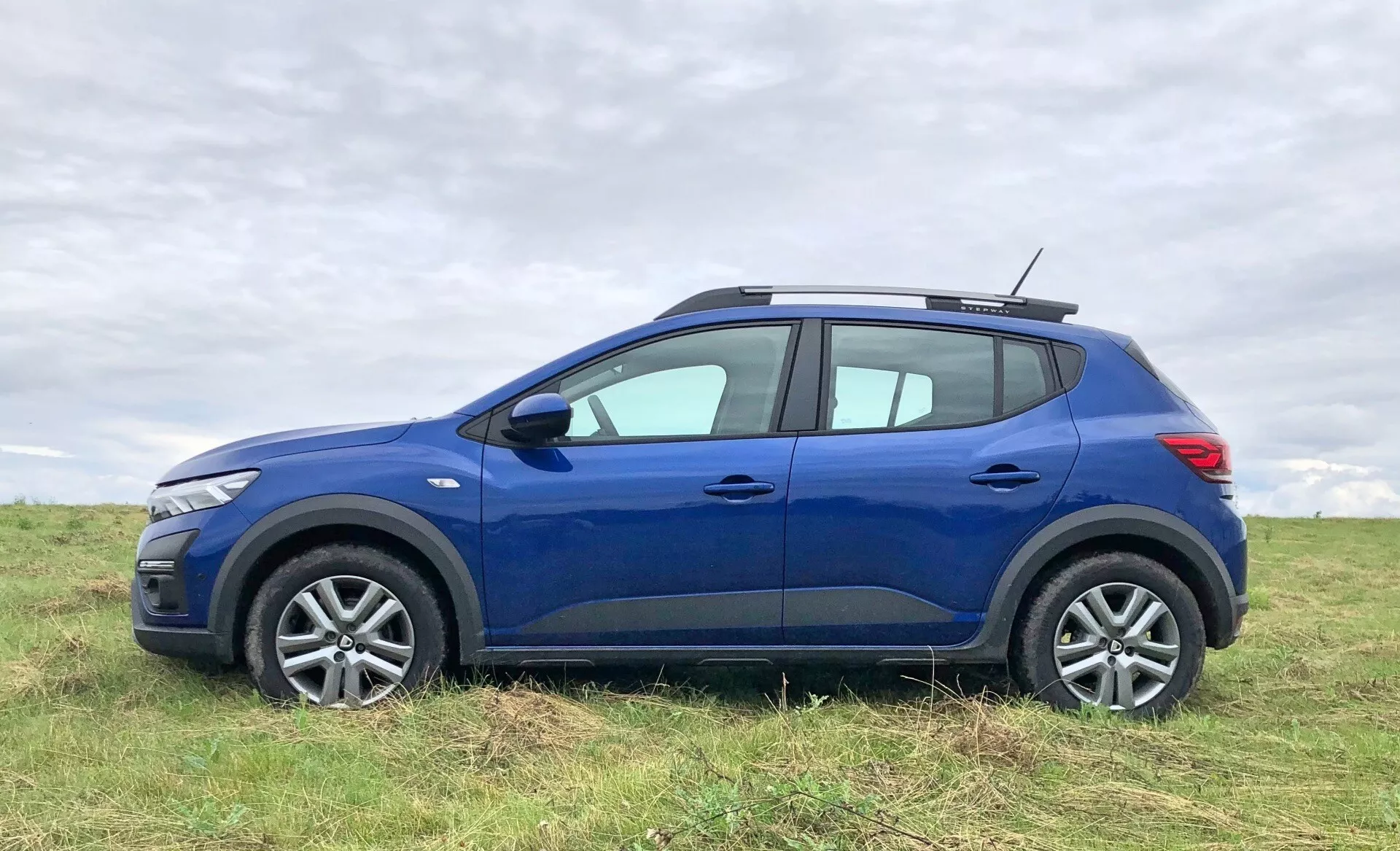 Obrázek modrého 2020 Dacia Sandero Stepway, crossover SUV, zaparkovaného na otevřeném poli. Auto je zobrazeno z bočního profilu, zvýrazňujícího moderní design předního světla, litá kola a strukturované pneumatiky. Exteriérové prvky zahrnují zpětná zrcátka, kliky dveří, charakterové linie a mírný oblouk nad blatníky. Linie střechy se mírně svažuje směrem k zadní části a obsahuje střešní nosič, který přidává na užitkové hodnotě. Viděný je také zadní světlomet a zatmavená skla na zadních oknech. Pozadí ukazuje jemně rozmazanou zamračenou oblohu a trávu, což činí vůz hlavním objektem snímku.