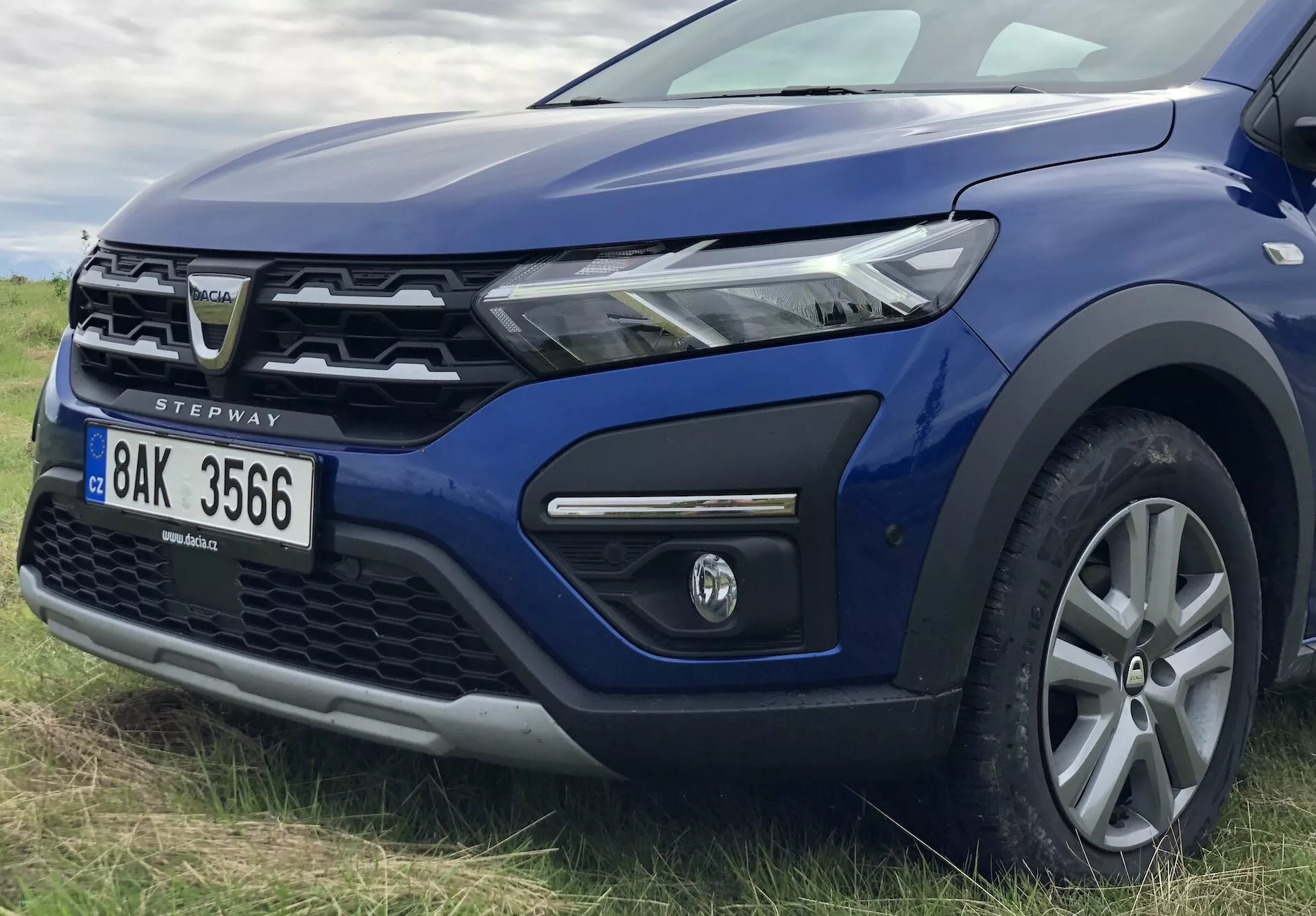 Detailní fotografie modrého Dacia Sandero Stepway 2020 z předního levého čtvrtletí, která zobrazuje moderní LED světlomet, částečně viditelnou masku s charakteristickým logem, přední nárazník s mlhovkou v černém rámu, zvýšenou kapotu, litinové kolo s několika paprsky, zrcátko ve stejné barvě jako karoserie, plastové obložení na oblouku kola a dveří naznačující schopnost jízdy v terénu, skloněné čelní sklo a A-sloupek a odznak 'STEPWAY' označující model.