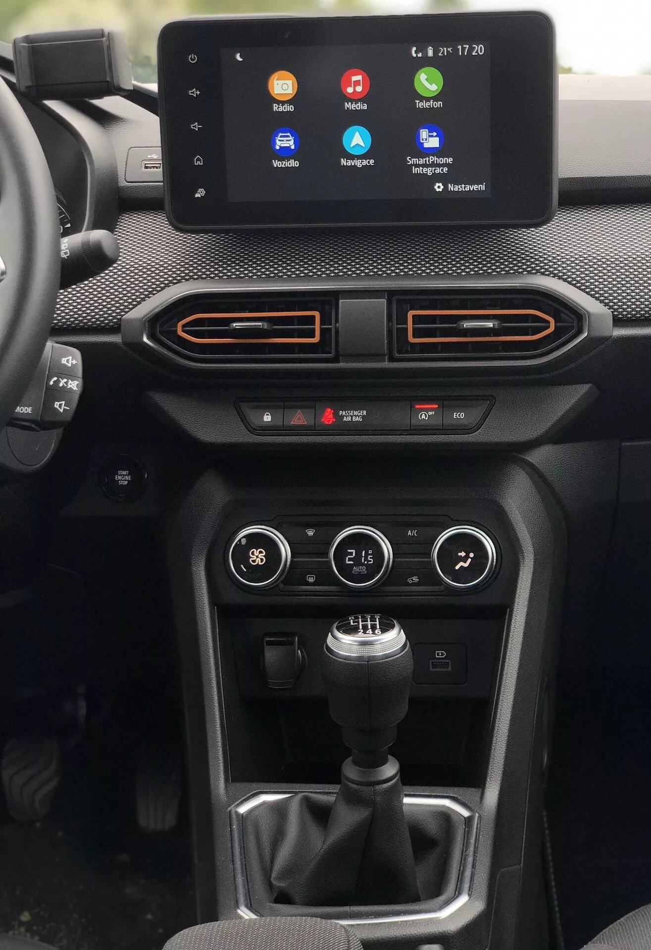 Pohled do interiéru Dacia Sandero Stepway 2020, zaměřený na středovou konzoli z pohledu řidiče. Významné prvky zahrnují infotainmentový dotykový displej zobrazující barevné ikony aplikací pro hovory, navigaci a streamování hudby, s časem a teplotou v pravém horním rohu. Pod obrazovkou jsou dva horizontální průduchy s oranžovými akcenty, panel klimatizace s tlačítky a otočnými ovladači a manuální řadicí páka na texturovaném podkladu. Tlačítko vpravo naznačuje různé jízdní režimy nebo ovladače a pod ovládáním klimatizace jsou viditelné dva porty USB. Materiály zahrnují kombinaci měkkého dotykového přístrojového panelu, syntetických nebo textilních textur a plastů. Navíc je zde ambientní osvětlení kolem okrajů ventilačních panelů odpovídající oranžové barvě. Kompozice nabízí jasný pohled bez zkreslení, mírně nakloněná doprava pro vyvážené zobrazení.