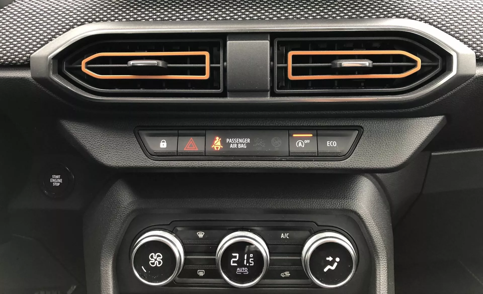 Detailní pohled na interiér vozu Dacia Sandero 2020 (Sandero-Stepway), zaměřený na středový panel. Obrázek zobrazuje dvě horizontálně orientované větrací otvory s kartáčovaným kovovým povrchem a oranžovým lemováním, tlačítko nouzových světel, indikátor stavu airbagu spolujezdce, indikátor zádržného systému vozidla, tlačítko ekologického režimu jízdy, tlačítko rozmrazování a klimatizační systém se třemi otočnými knoflíky, které ukazují rychlost ventilátoru, teplotu (21,5 °C) a směr proudění vzduchu. Další detaily zahrnují texturu podobnou uhlíkovým vláknům a černé matné materiály, podsvícený displej a podsvícená tlačítka.