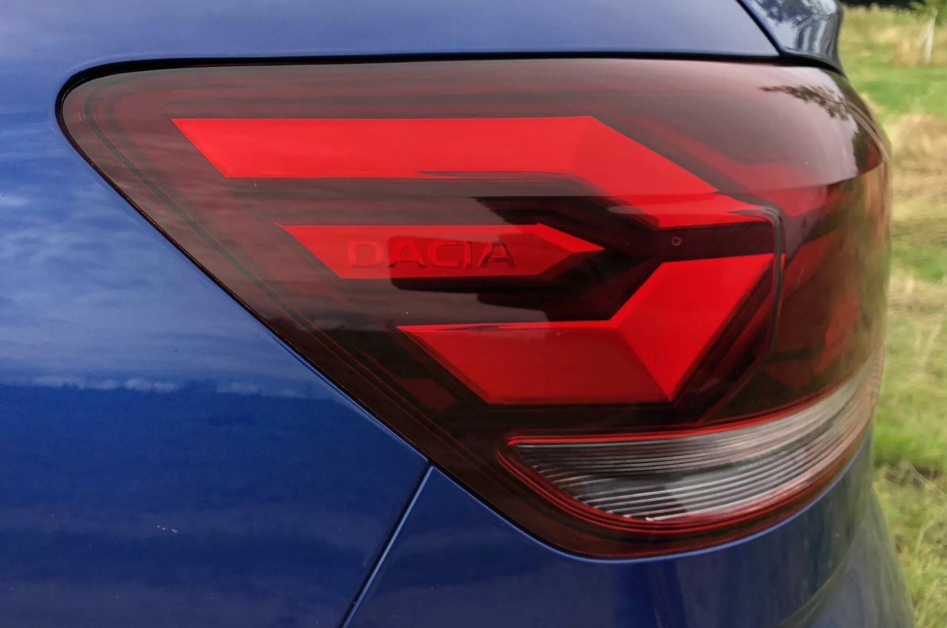 Detailní pohled na zadní levou stranu Dacia Sandero Stepway 2020 se zaměřením na moderní design zadního světla. Zadní světlo s úhly a červeným LED vzorem ve tvaru chevronu naznačuje pohyb vpřed. Jasná sekce pravděpodobně obsahuje zpáteční světlo a směrovku. Slovo 'DACIA' je viditelné v obalu světla. Nad zadním světlem je částečně viditelný modrý lak karoserie.