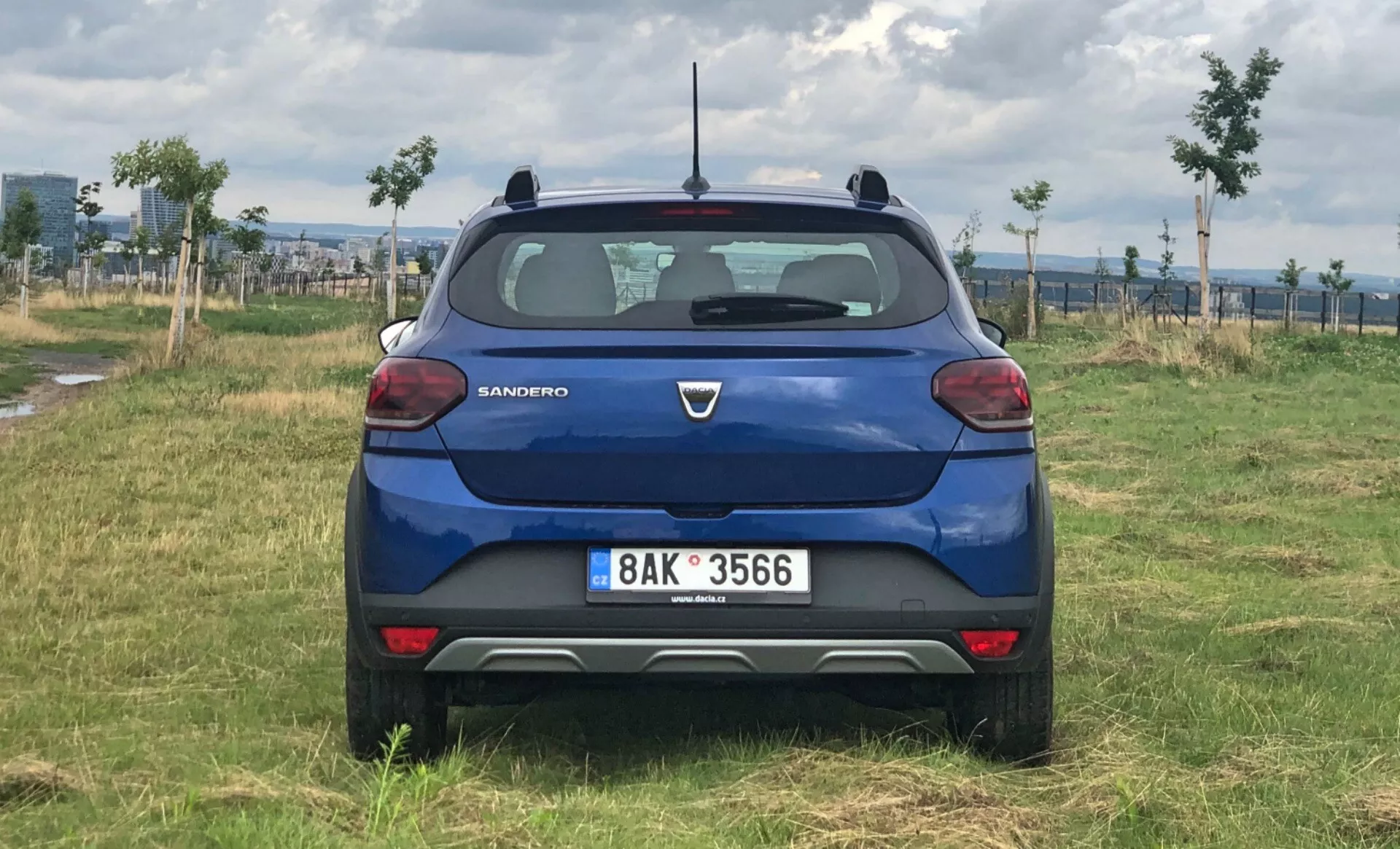 Pohled zezadu na modrý hatchback Dacia Sandero 2020 na nezpevněném travnatém terénu s stromy a možná vinicí v pozadí. Auto je kompletně v záběru, ukazuje obě zadní světla, registrační značku, zadní čelní sklo a víko kufru s logem výrobce.