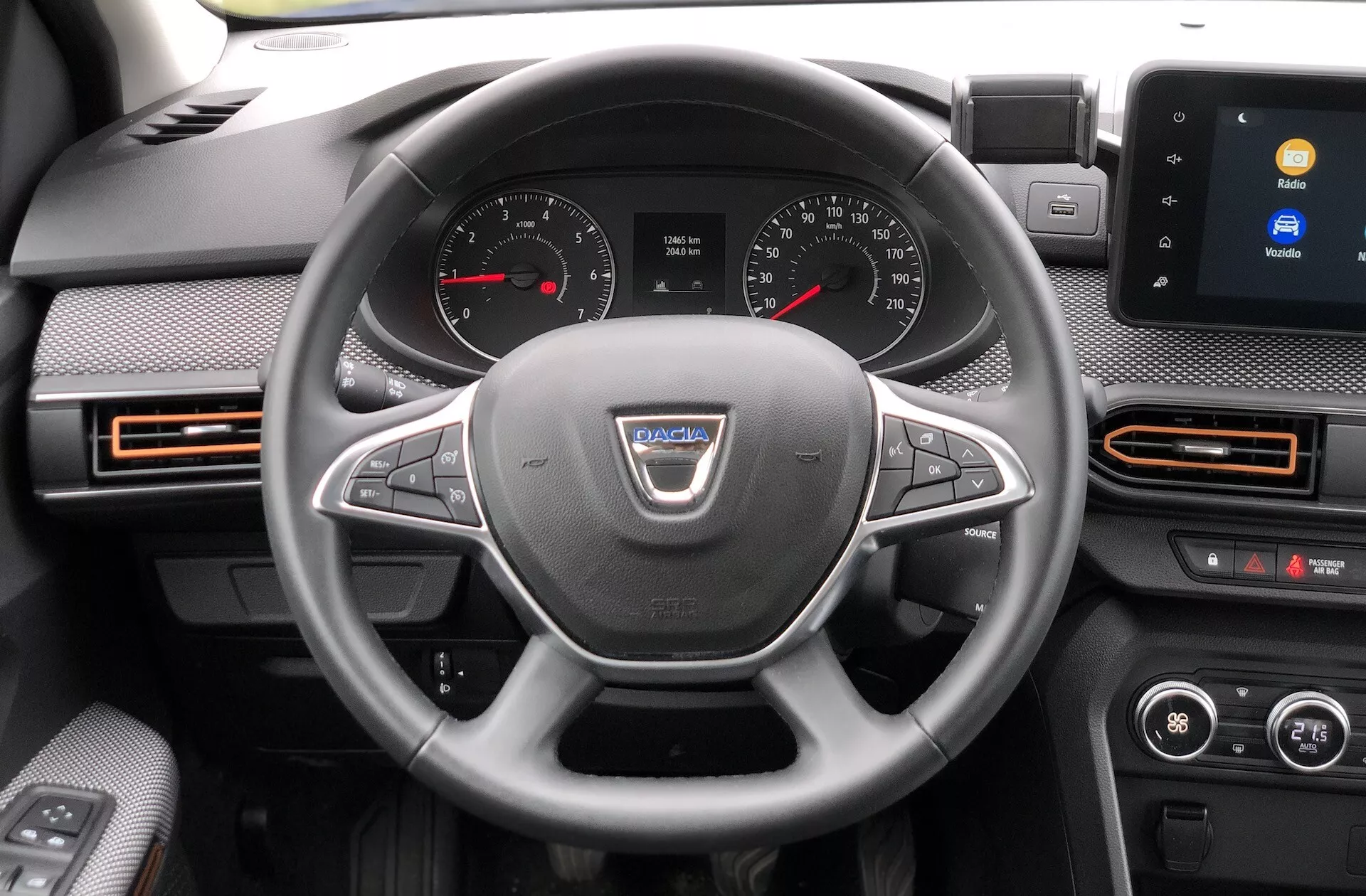 Pohled do interiéru Dacia Sandero Stepway 2020 zaměřený na palubní desku z pohledu řidiče. Volant, centrálně umístěný na fotografii, obsahuje logo výrobce a několik ovládacích tlačítek pro funkce jako ovládání hlasitosti, hands-free volání a tempomat. Za volantem je vidět přístrojový panel s analogovými ciferníky pro tachometr a otáčkoměr a digitálním displejem mezi nimi. Uprostřed palubní desky se nachází dotykový infotainment systém zobrazující možnosti připojení, jako je Bluetooth. Viditelné jsou také otvory s oranžovými akcenty, dvouzónová klimatizace a náznaky dveří řidiče s elektricky ovládanými tlačítky oken a nastavením zpětných zrcátek.