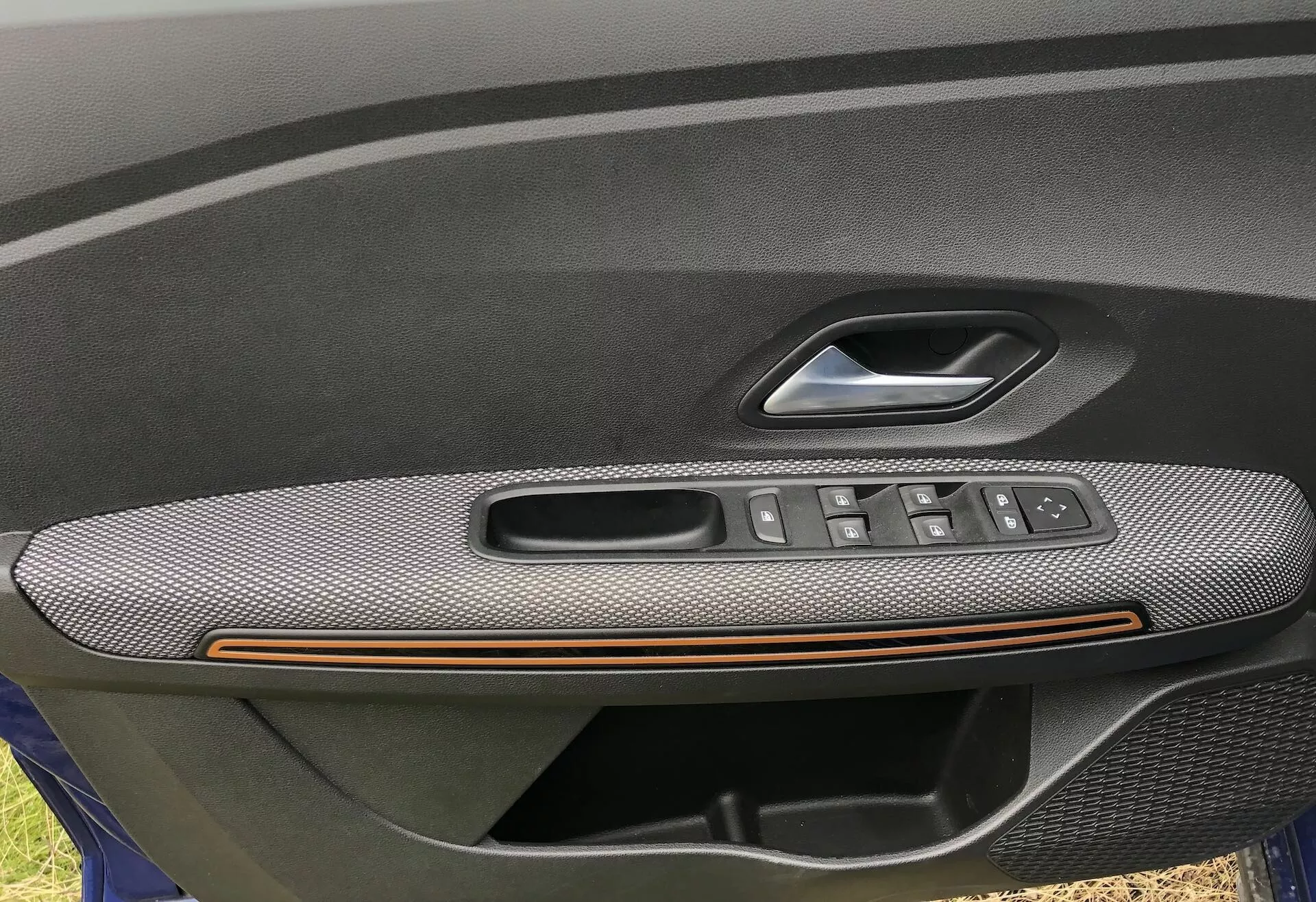 Detailní záběr na vnitřní panel dveří řidiče Dacia Sandero 2020 Stepway, který ukazuje opěrku s texturou uhlíkových vláken, bronzovým akcentním pruhem a integrovaným ovládacím panelem s tlačítky na elektrická okna, ovládacím joystikem na nastavení zpětných zrcátek a klikou dveří s tlačítkem pro odemknutí/zamknutí. Obrázek zdůrazňuje luxusní a praktické prvky, což naznačuje vyšší úroveň výbavy.