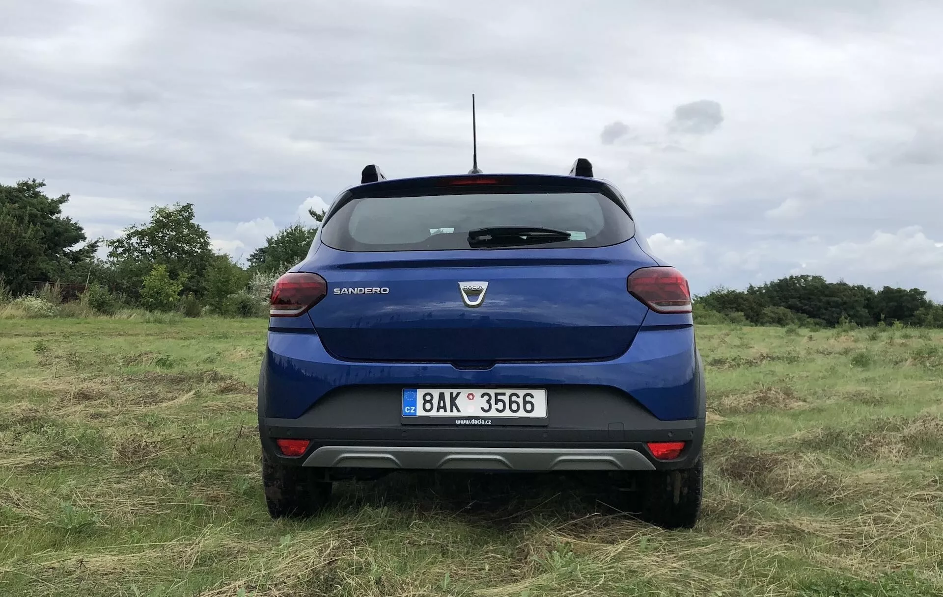 Pohled zezadu na modrou Dacii Sandero Stepway 2020 zaparkovanou na nerovném travnatém poli, se značkou a názvem modelu viditelně zobrazenými na zadních dveřích. Zadní sklo vozidla má jediný stěrač, na bocích jsou viditelné zadní světla s červenobílým barevným schématem. Viditelné jsou vysoké brzdové světlo u základny zadního okna, střešní anténa a dvoubarevný zadní difuzor. Nárazník má možné parkovací senzory, SPZ je centrálně pod logem značky a prohlubeň může sloužit jako prostor pro kameru.