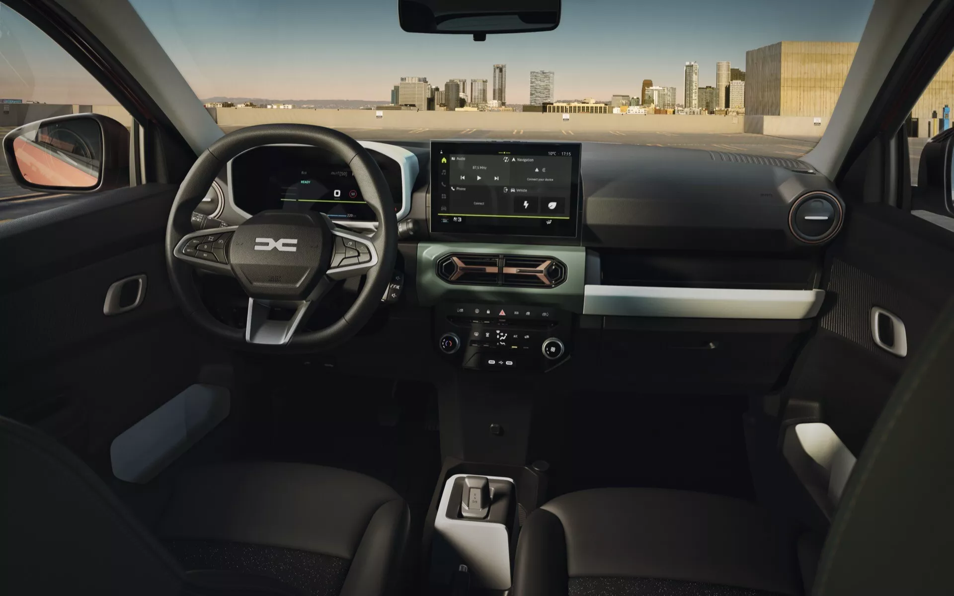 Interiér Dacia Spring facelift 2024 z pohledu zadního sedadla ukazující volant s ovládacími tlačítky, digitální palubní desku, dotykovou obrazovku infotainment systému, systém klimatizace a páku automatické převodovky