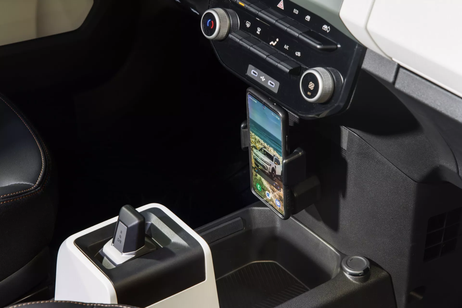 Detailní pohled na středovou konzoli modelu Dacia Spring 2024 po faceliftu s pákou řazení, ovládáním klimatizace a částečně zobrazeným displejem infotainmentu s držákem pro smartphone pro navigaci GPS, zdůrazňující moderní design interiéru ve vyspělém technologickém vybavení vozidla