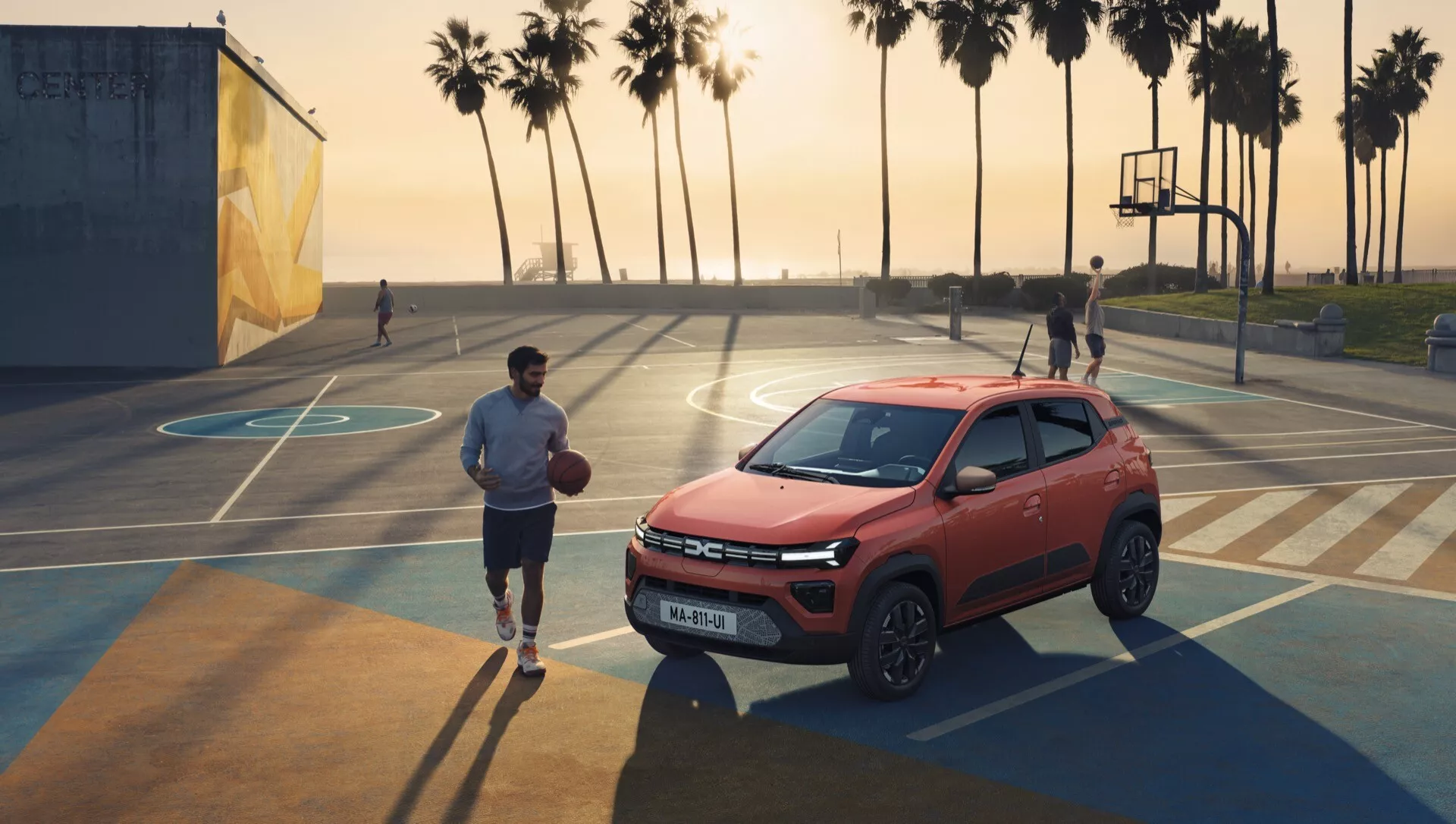Facelift Dacia Spring 2024, kompaktní SUV ve vysokém pohledu zepředu a z boku na basketbalovém hřišti, ukazuje pokročilé osvětlení, mřížku s čidly, slitinová kola a praktický a přitom sportovní design naznačující terénní schopnosti