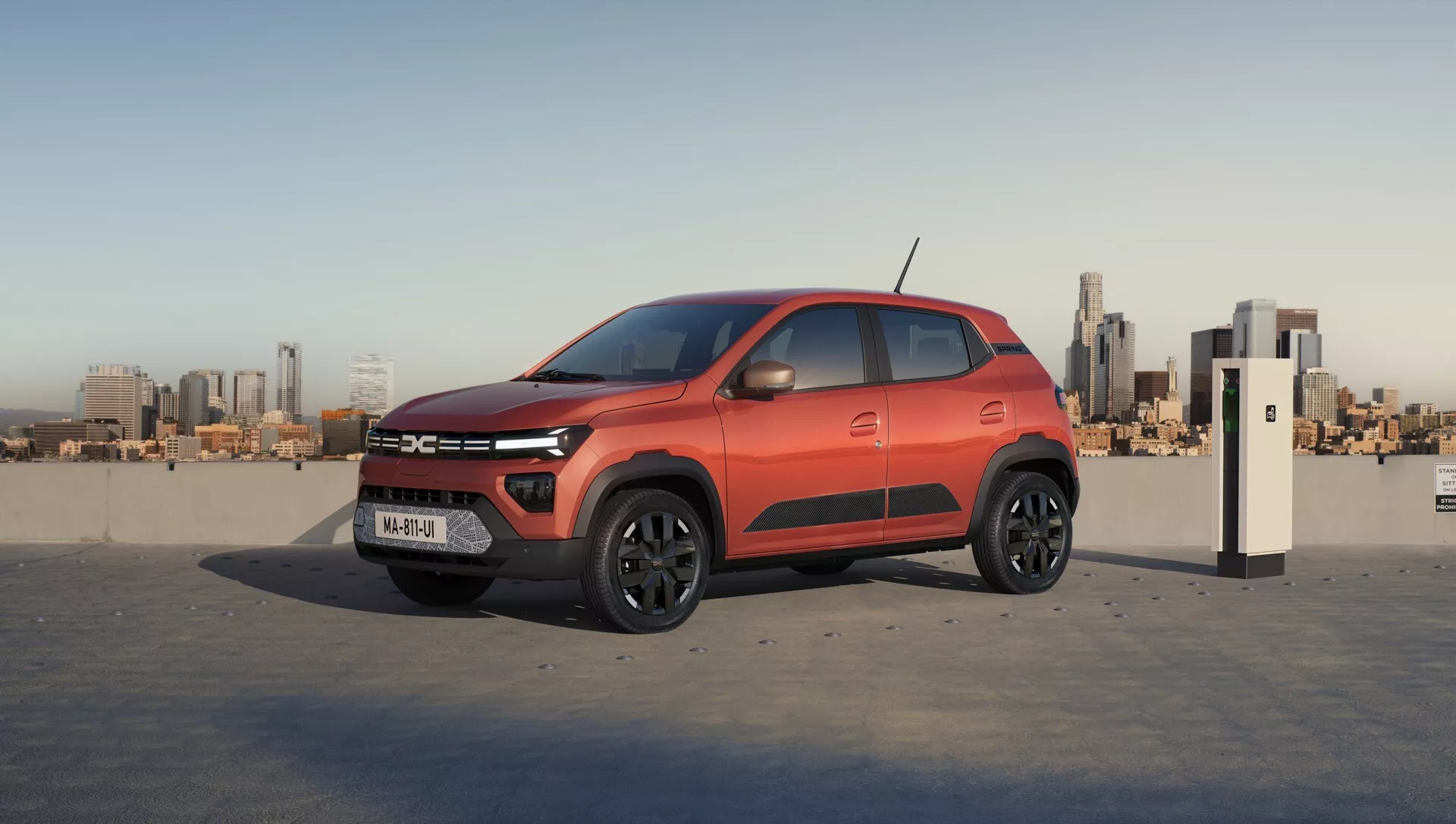 Facelift první generace Dacia Spring 2024 - Moderní kompaktní SUV s výrazným červeno-černým dvoubarevným schématem, štíhlými LED světlomety, slitinovými koly a pokročilou technologií, zaparkované proti městskému panoramatu