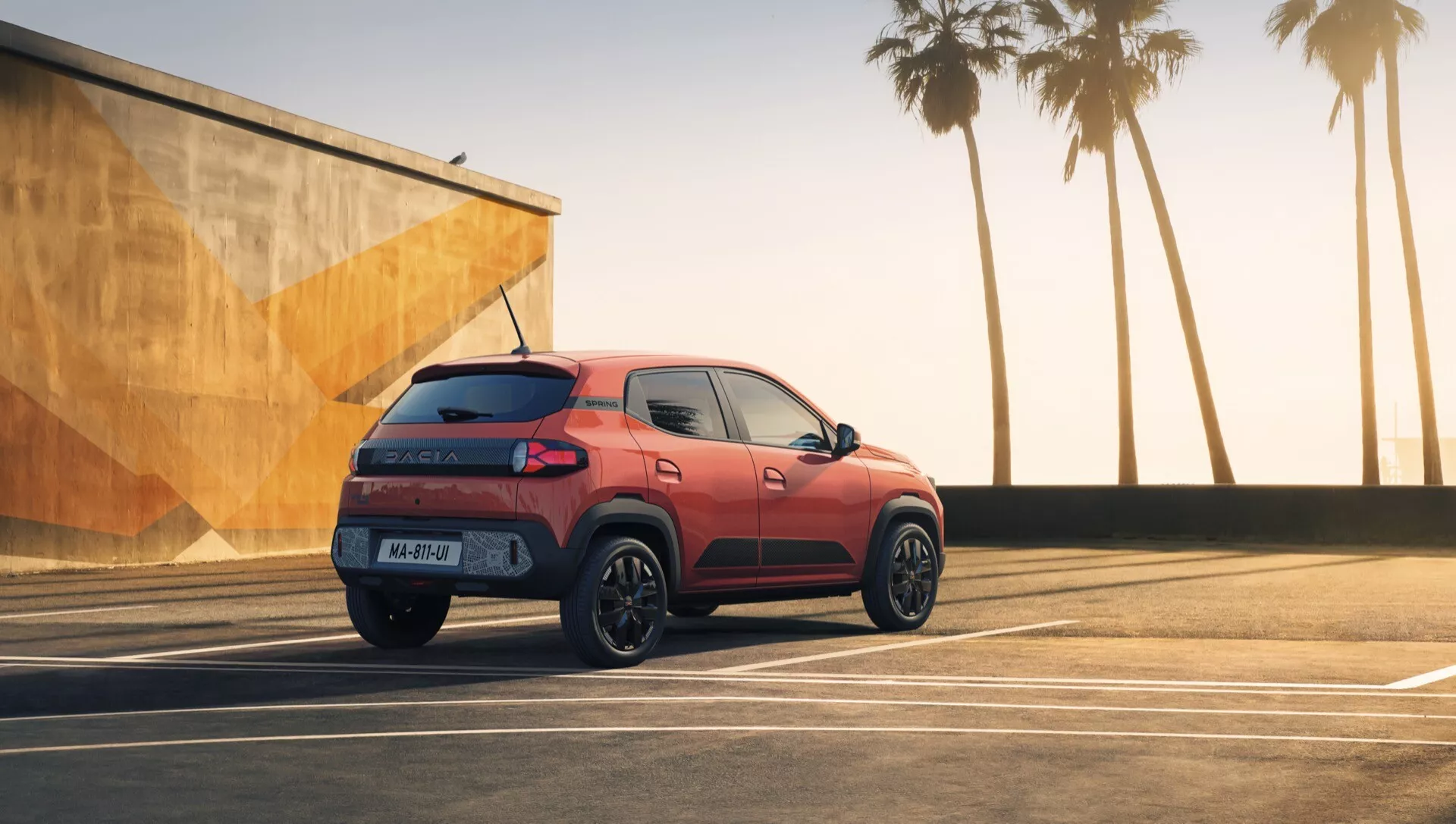 Facelift model kompaktního SUV Dacia Spring 2024 zaparkovaný na prostorném parkovišti za soumraku, zdůrazňující jeho zadní tříčtvrteční pohled, zadní LED světla, sportovní černá kola a aerodynamický design na pozadí pobřežního městského prostředí
