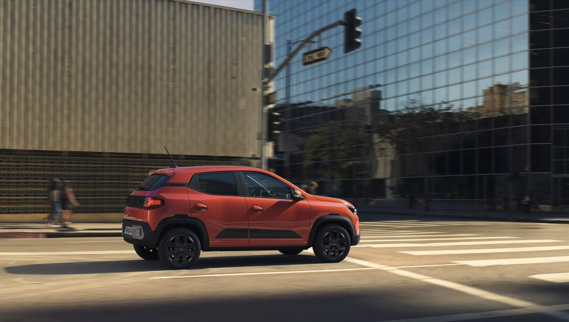 Oranžové kompaktní SUV Dacia Spring 2024 (1. generace - facelift) v dynamickém bočním profilu ukazující sportovní design a postoj na silnici, v pohybu s rozmazáním pohybu, na pozadí městského prostředí ve dnešním denním světle