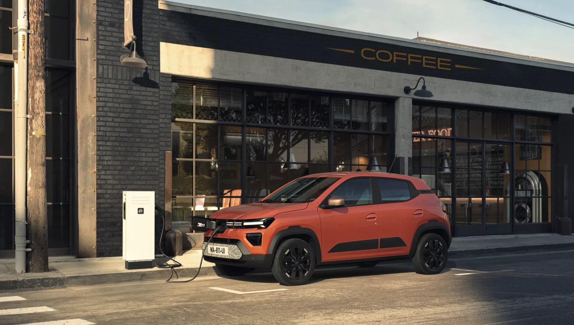 Facelift Dacia Spring 2024 - Moderní elektrické SUV v červené s černými akcenty, nabíjí na stanici, boční pohled se zaměřením na zadní polovinu, městské prostředí