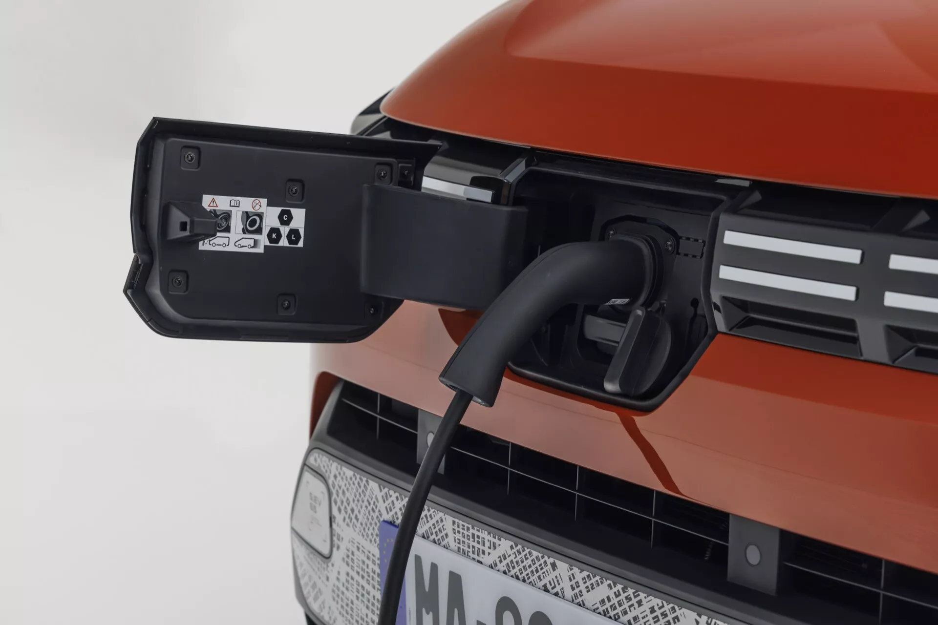 Detailní záběr na nabíjecí port elektrického vozidla Dacia Spring 2024 v jasně oranžové barvě s připojeným kabelem, s černou mřížkou naznačující možnosti autonomního řízení a pokročilým rozhraním konektoru