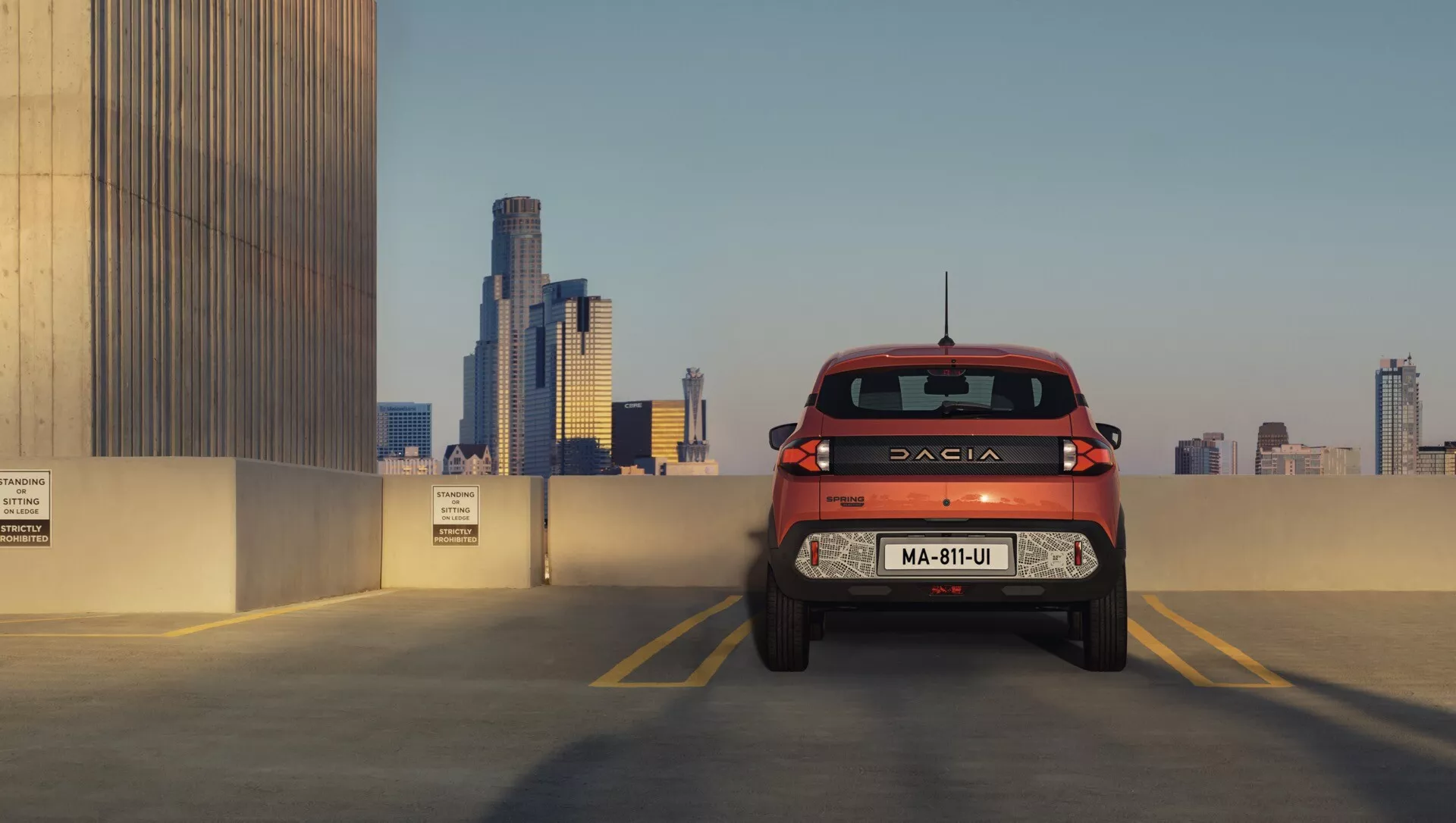 Facelift Dacia Spring 2024: Detailní pohled na červenou Dacii z úhlu střední zadní části s důrazem na zadek vozu, design segmentových zadních světel a značení na parkovišti na střeše s městským panoramatem ve dne