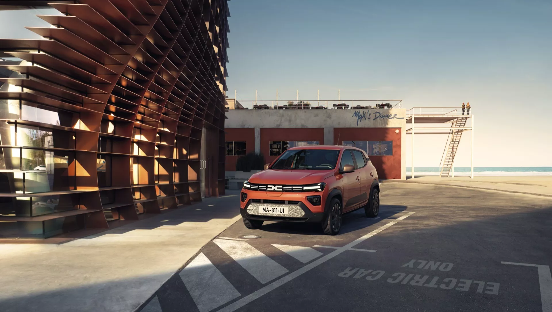 Dacia Spring 2024 v první generaci po faceliftu zaparkovaná na malebném střešním parkovišti nebo v přímořské oblasti, předvádějící její kompaktní SUV design, LED světlomety, denní svícení a slitinová kola na pozadí klidného oceánu pod čistou oblohou