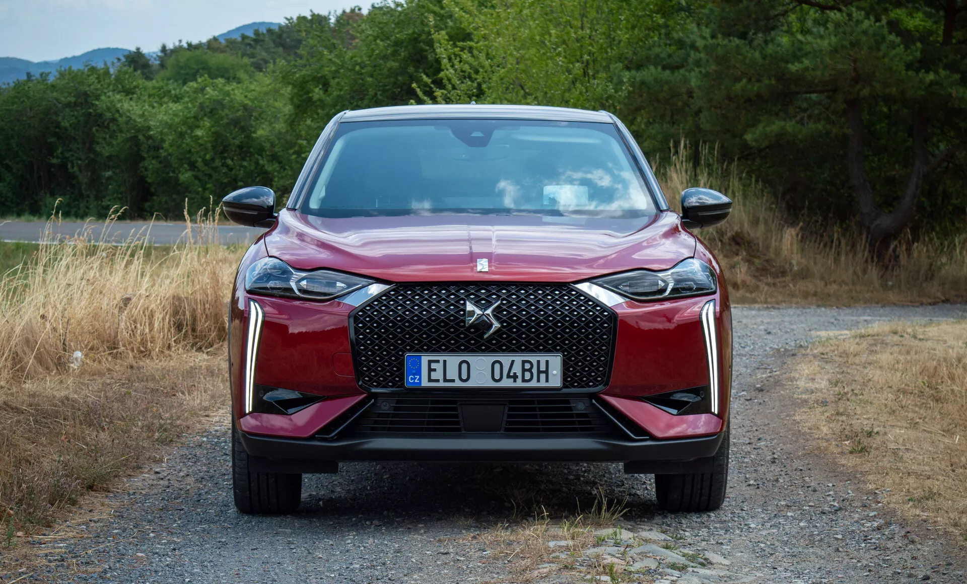 Frontální pohled na DS Automobiles DS 3 E-TENSE z roku 2023 zaparkovaný na štěrkové cestě s zelení v pozadí, představující přední mřížku, LED světlomety, linie kapoty a design nárazníku