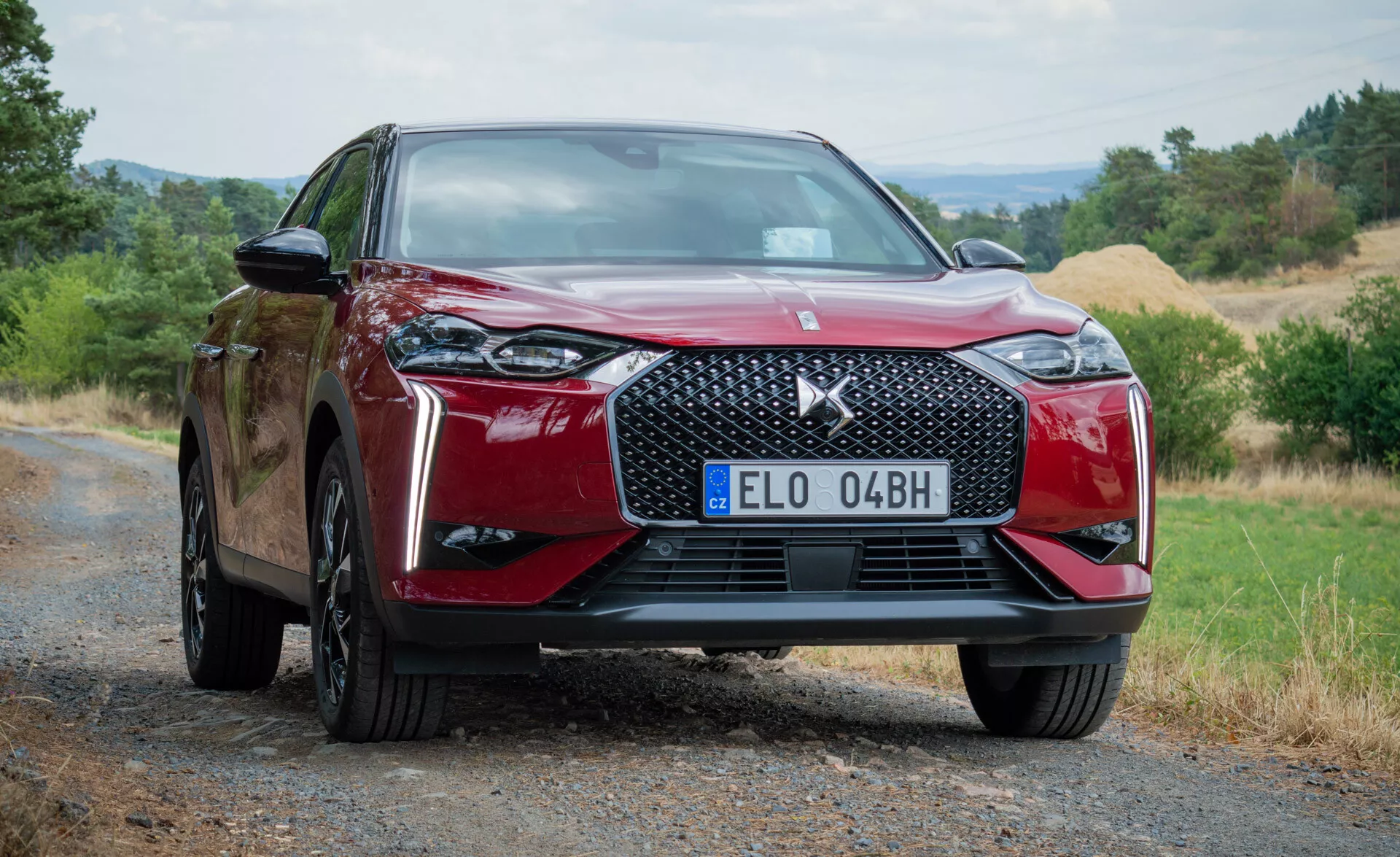 Moderní červené vozidlo DS Automobiles DS 3 E-TENSE 2023 z nízkého předního úhlu zdůrazňující přední mřížku, emblém a částečný boční profil na štěrkové cestě s přírodním pozadím