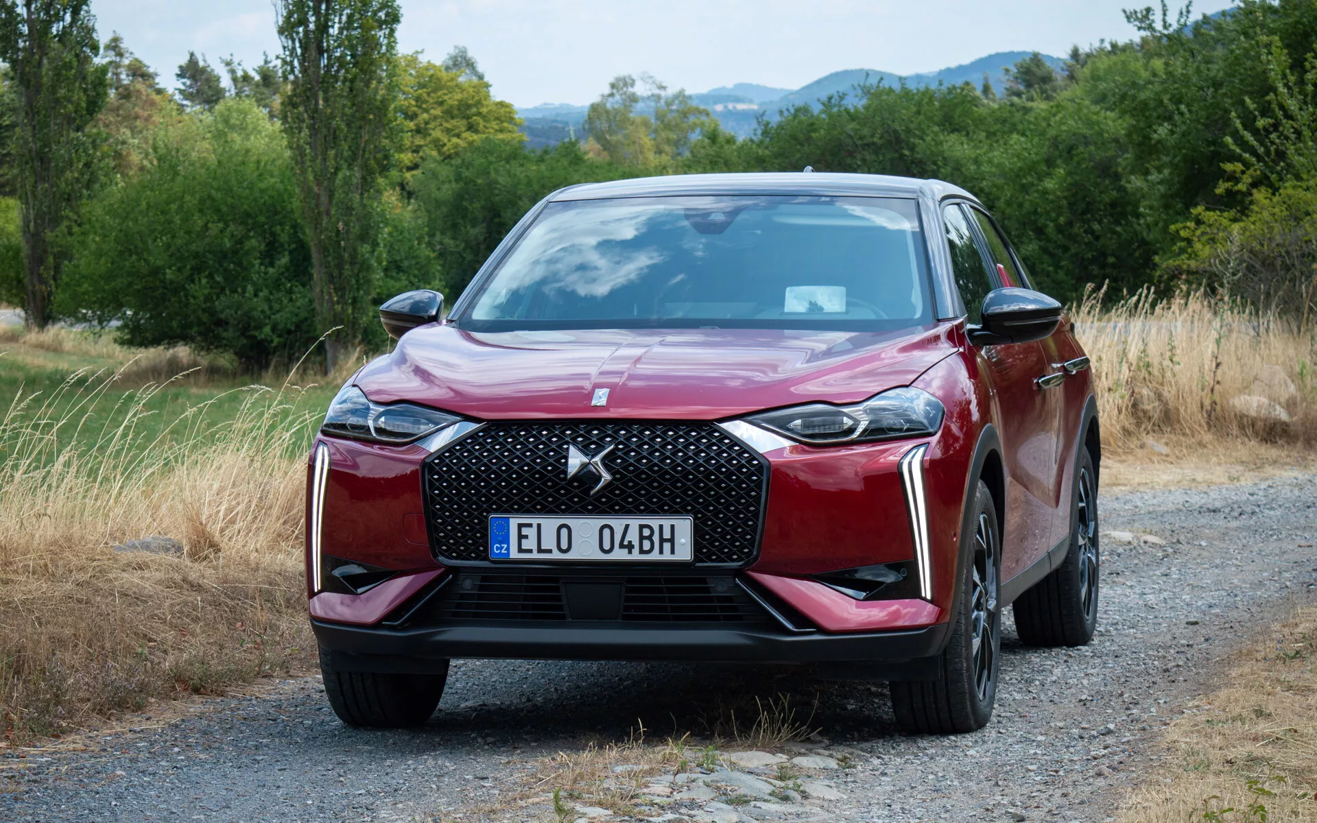DS Automobiles DS 3 E-TENSE 2023 zobrazený na štěrkové cestě, ukazuje svou šestihrannou mřížku, LED světlomety, sofistikovaný přední nárazník a metalický červený lak na pozadí přírodní scenérie se stromy a trávou