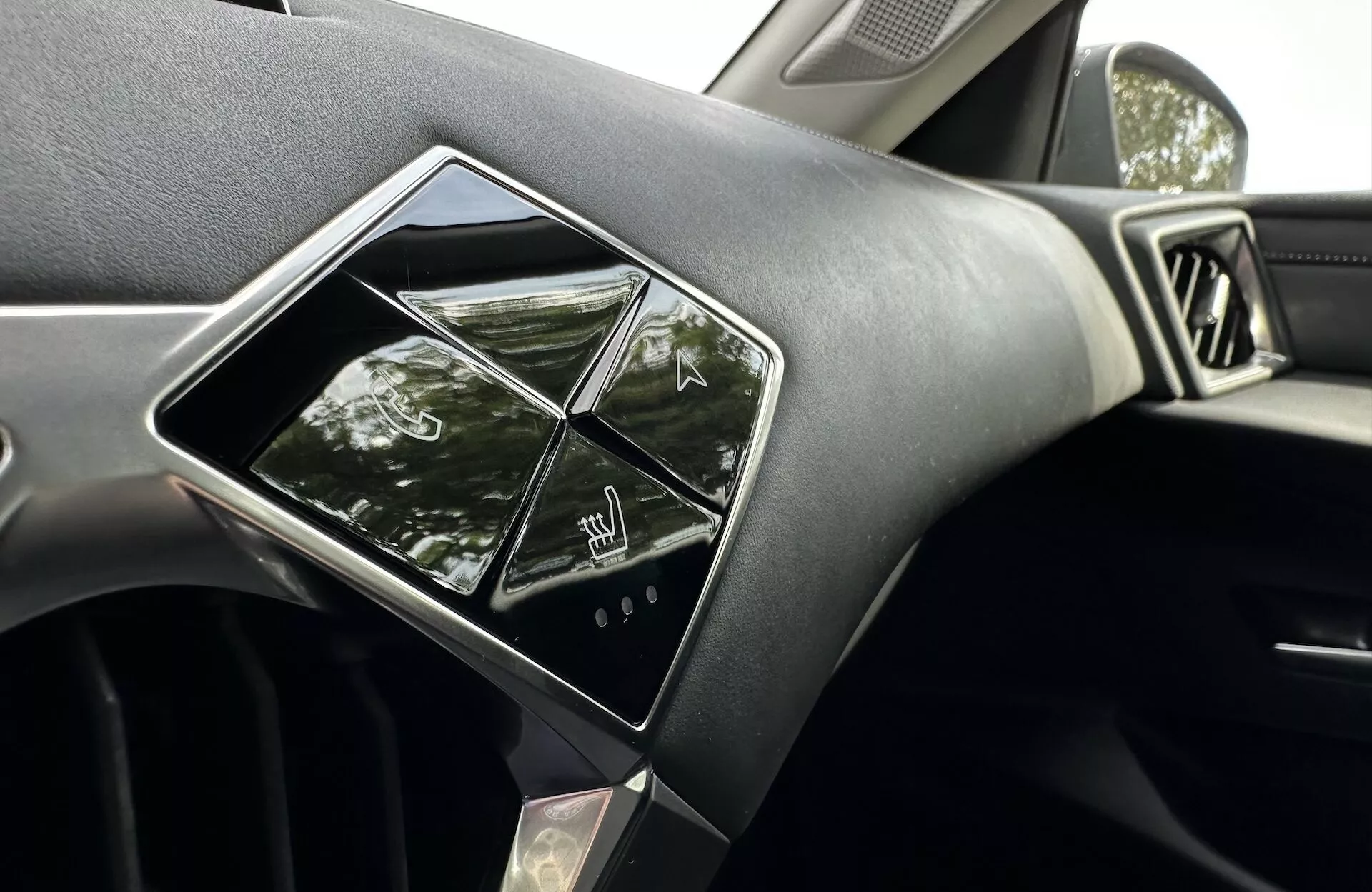 Vnitřní pohled na DS 3 E-TENSE 2023 se zaměřením na palubní desku a systém infotainment, obsahující dotykově citlivé ovladače, prémiový design palubní desky s materiálem připomínajícím měkký dotek nebo kůži, část ventilační mřížky a odrazy naznačující přirozené denní světlo a venkovní parkovací prostředí.