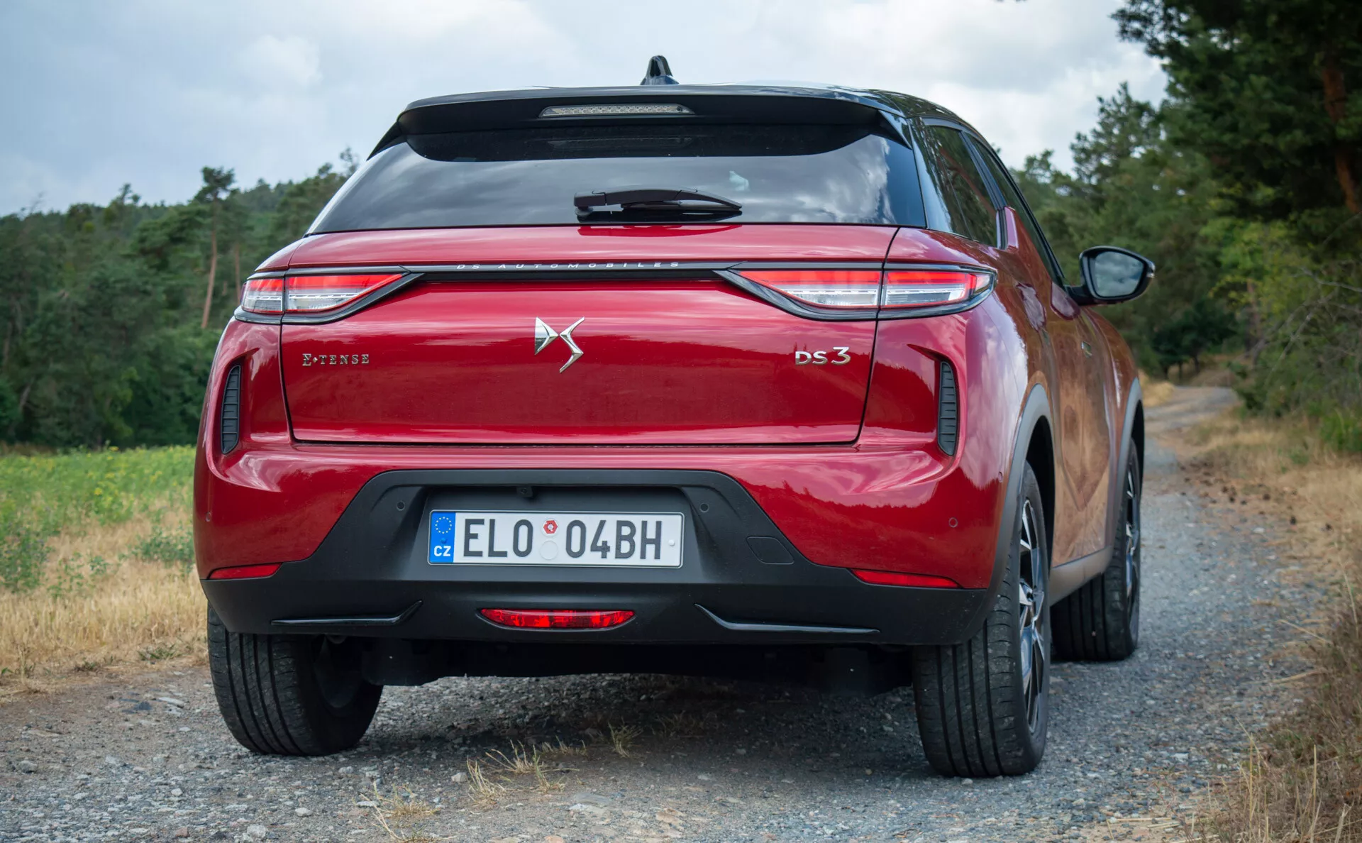 Zadní pohled na červený model DS 3 E-TENSE 2023, předvádějící moderní konstrukci zadních světel, dvojitých výfuků a označení, umístěný venku na štěrkové silnici s vegetací.