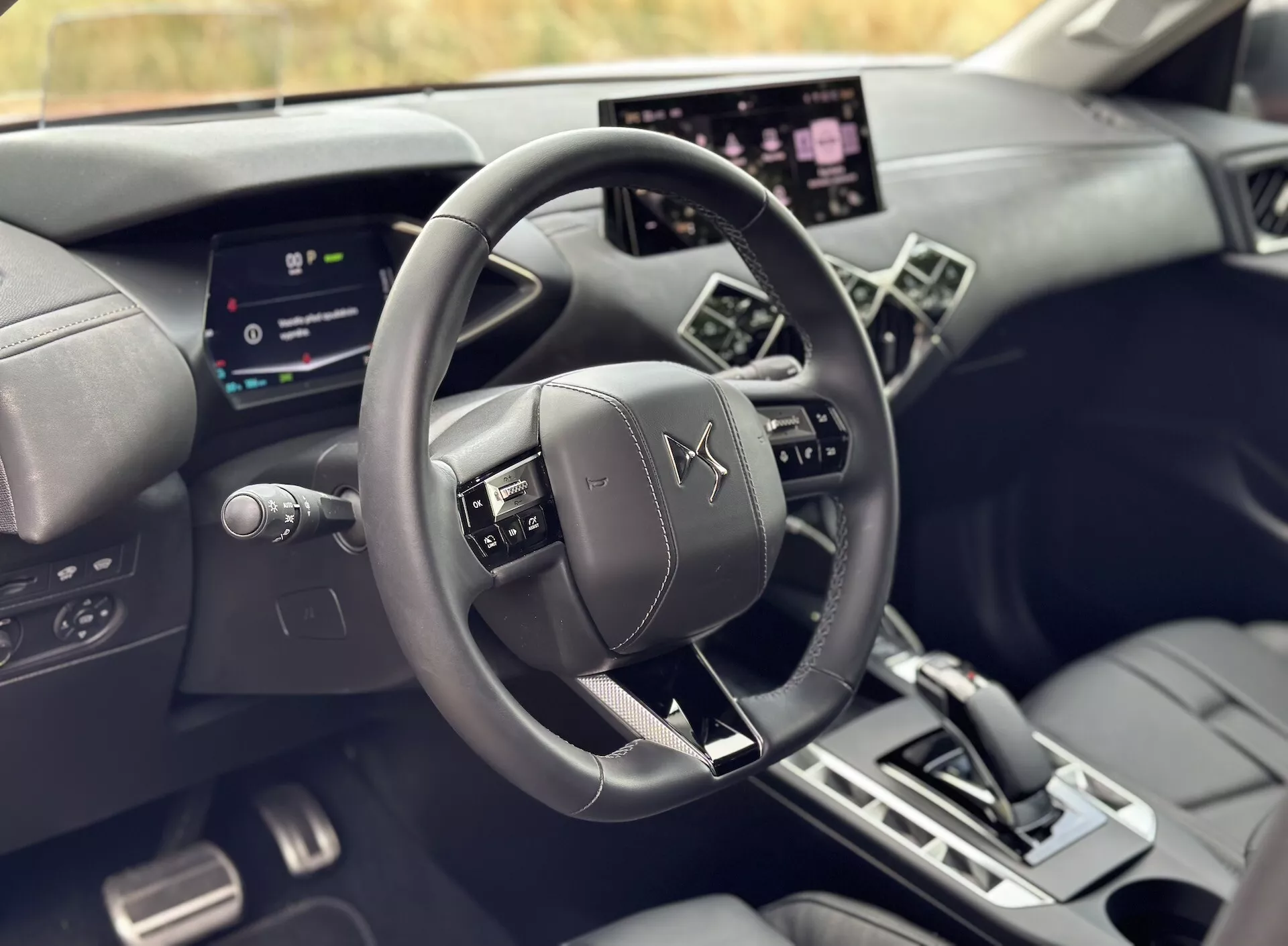 Interiérový pohled z perspektivy řidiče na DS 3 E-TENSE 2023 s centrálním kůží potaženým volantem s emblémem DS Automobiles, digitálním přístrojovým panelem a velkým dotykovým informačním systémem mezi luxusními tmavými tóny s angulárními designovými liniemi.