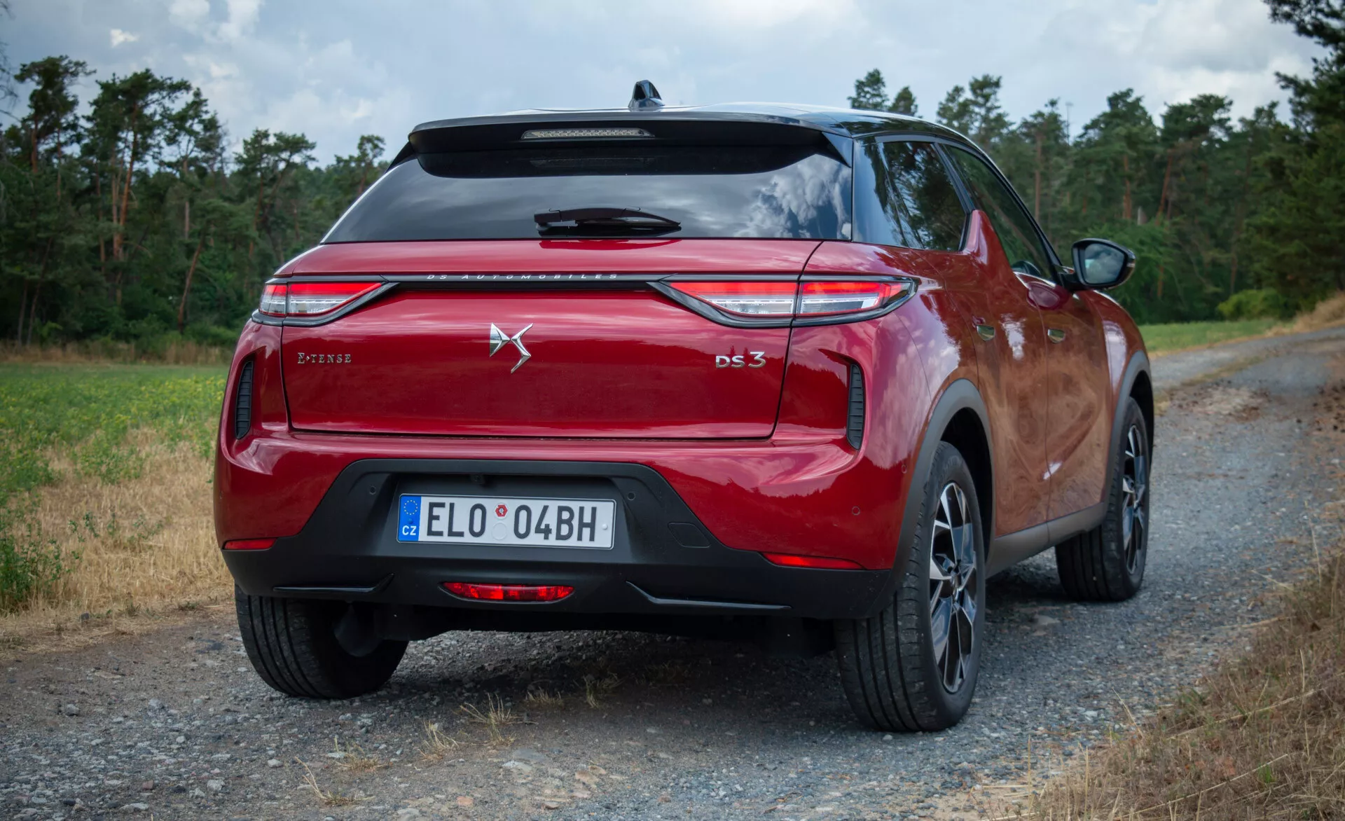 Hatchback DS Automobiles DS 3 E-Tense 2023 v metalické červené barvě, zobrazen ze zadního tříčtvrtečního úhlu s důrazem na zadní světla, emblém a označení modelu, umístěný na prašné cestě s přírodním pozadím.