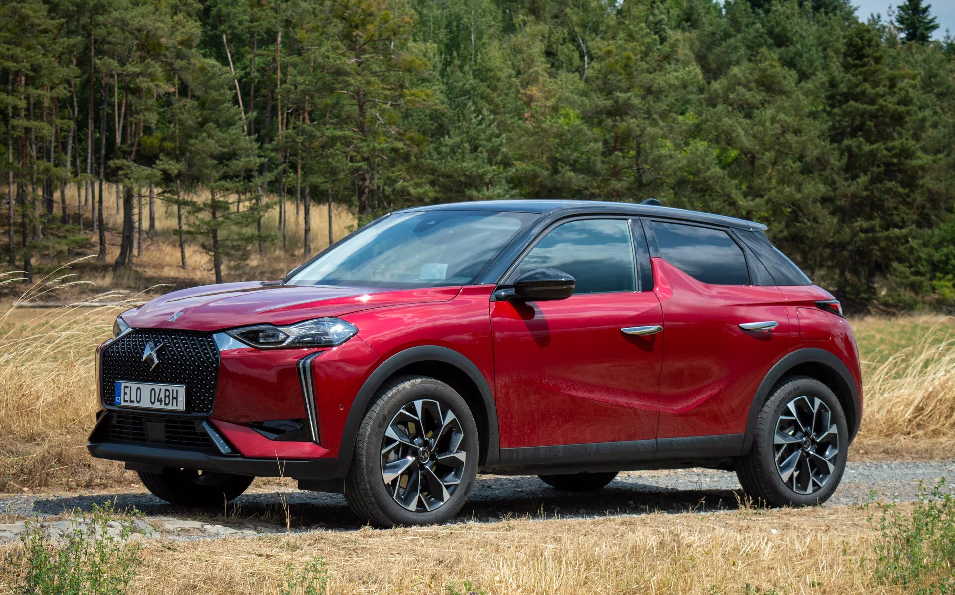 Boční přední pohled na DS Automobiles DS 3 E-TENSE 2023, červené SUV crossover, slitinová kola s více paprsky, LED světlomety, na štěrkové cestě s pozadím lesa