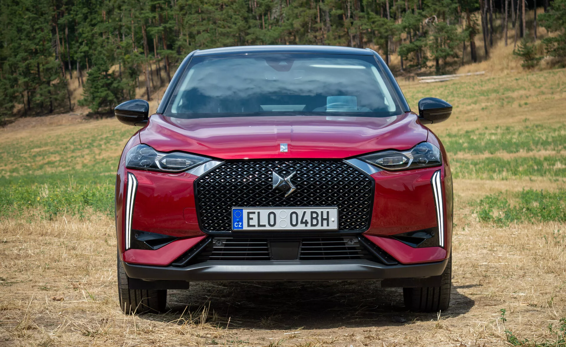 DS Automobiles DS 3 E-TENSE 2023 v červené barvě, prezentace charakteristických prvků z předního úhlu na nezpevněném terénu s pozadím lesa, osvětlení uprostřed dne zdůrazňující texturovanou mřížku, LED světlomety, aerodynamický nárazník se vzduchovými vstupy, evropskou registrační značku a lesklou lakovanou povrchovou úpravu.