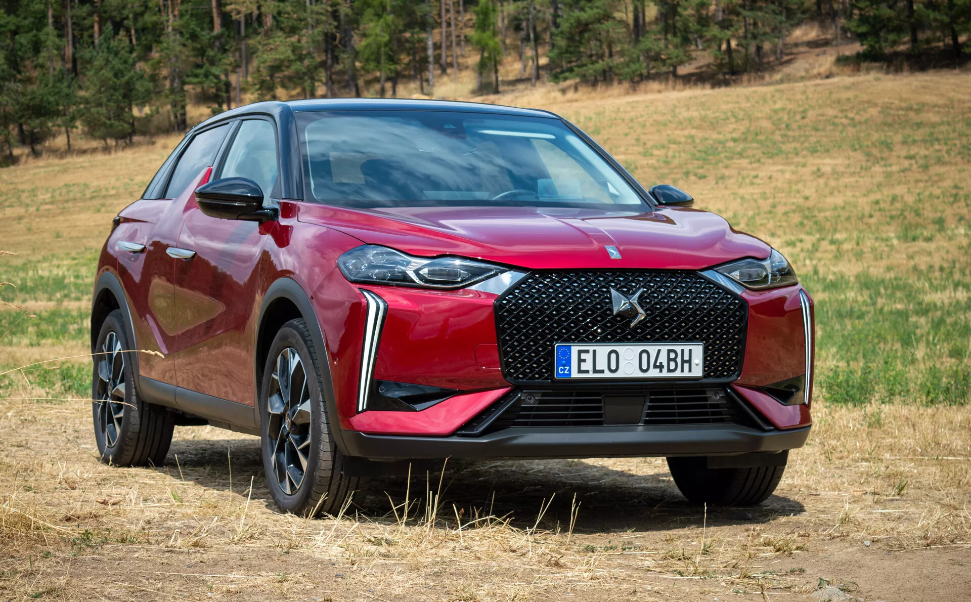Moderní SUV 2023 DS Automobiles DS-3 E-TENSE, pohled zepředu a ze strany ukazující mřížku, kapotu, světlomety a profil vozidla na suché trávě s přírodním pozadím