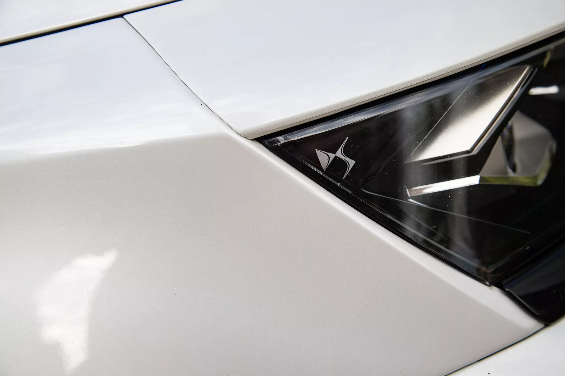 Detailní snímek světlometu DS Automobiles DS-7 2022 zobrazující místo, kde se blatník nebo kapota spojuje se světlometem, s lesklým, monochromatickým designem a značkovým emblémem uvnitř světlometu, což naznačuje moderní luxus a pokročilou technologii LED osvětlení.