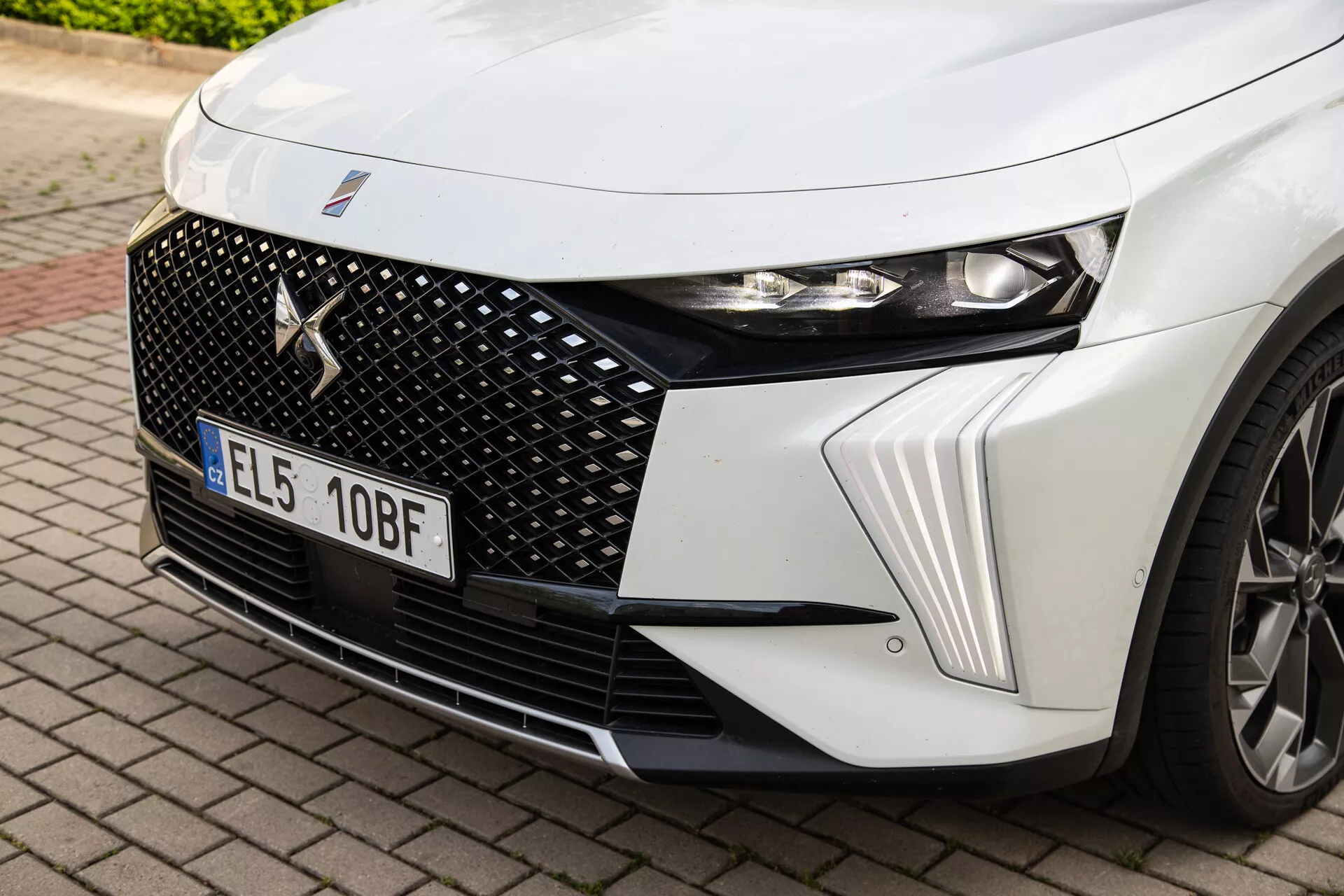 Detailní pohled na přední levý kvadrant DS Automobiles DS-7 2022 se zaměřením na LED světlomet, šestiúhelníkový design mřížky s centrálním odznakem značky a dynamické kontury kapoty spolu s náhledem na slitinové kolo