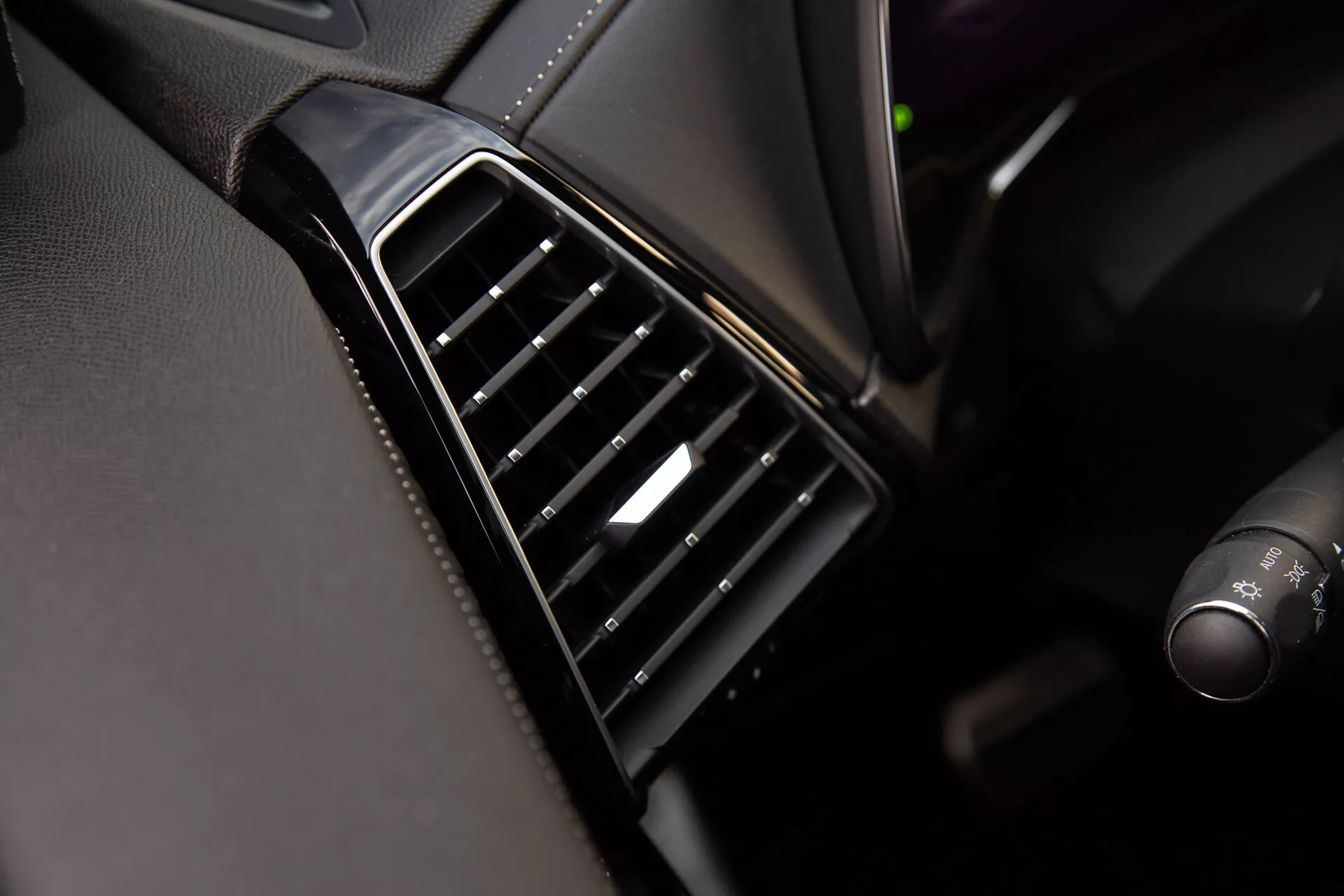 Detailní pohled na výdechový otvor v DS Automobiles DS-7 2022 na palubní desce s černými nastavitelnými lamelami uspořádanými vertikálně, lesklým rámečkem a stříbrným akcentem na ovladači, předvádějící kvalitu a materiály interiéru vozidla