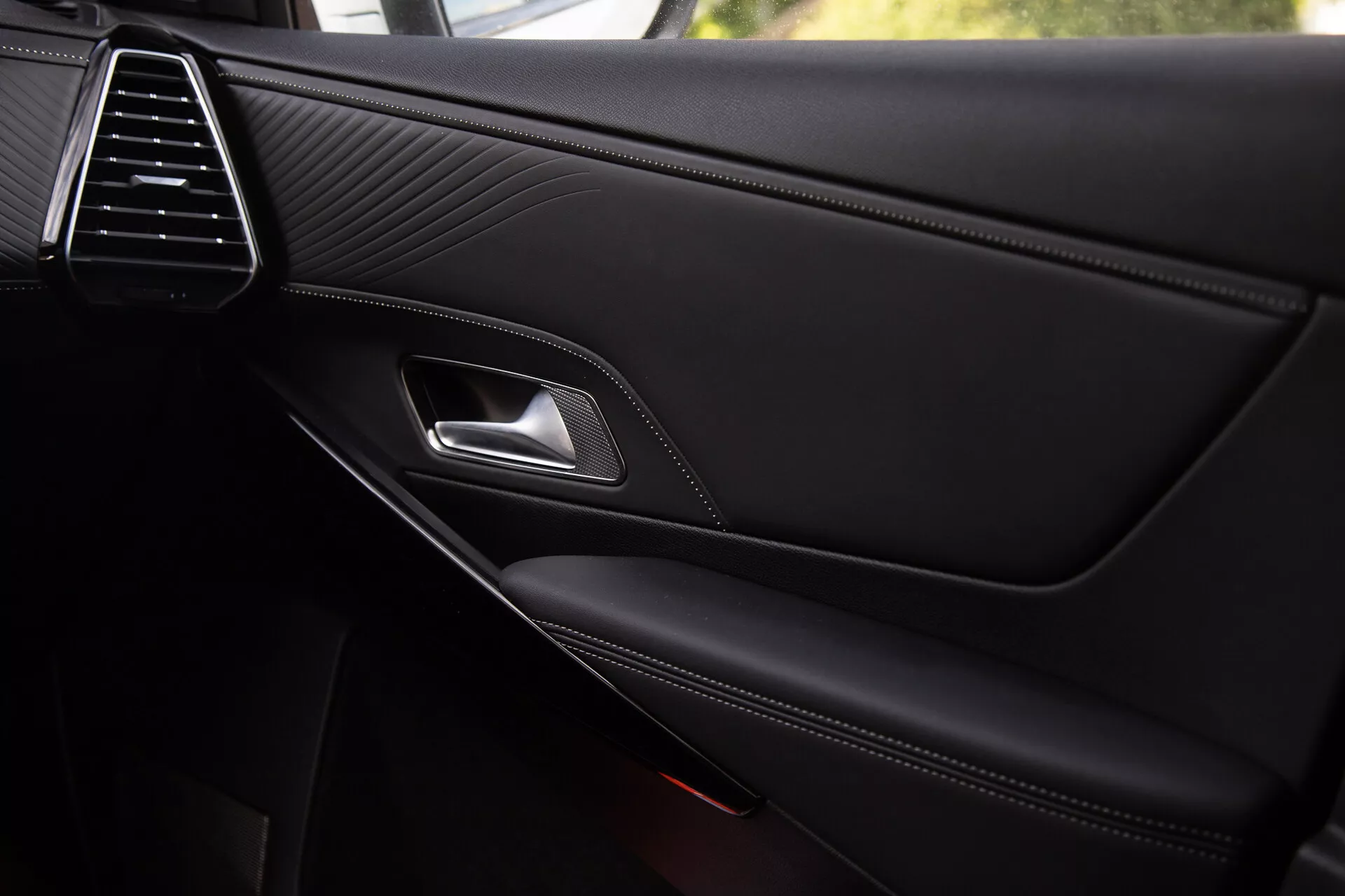 Detailní pohled na interiérový panel dveří DS Automobiles DS-7 2022 s elegantní větrací mřížkou, prošitou kůží, chromovanou kličkou dveří, ergonomickou páčkou zámku, kontrastně prošitou opěrkou ruky a integrovanou kapsou na drobnosti s částečně viditelnou mřížkou reproduktoru, což odráží prémiovou kvalitu a designové detaily.