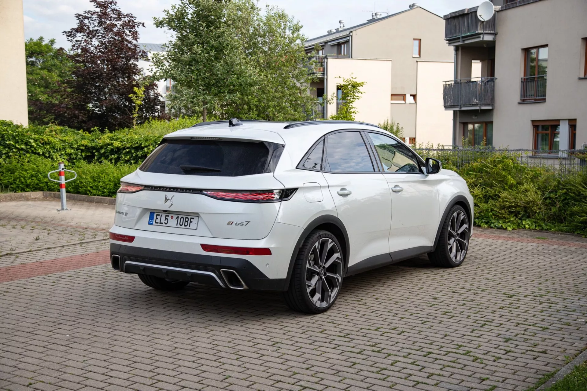 Bílé SUV DS Automobiles DS-7 ročník 2022 zaparkované na cihlově dlážděném prostoru, ukazující LED zadní světla, dvojitý výfukový systém a sportovní slitinová kola. Snímek zachycuje vozidlo s elegantním designem SUV podobným kupé z trojčtvrtě zadní perspektivy, s možným panoramatickým střešním oknem a zrcátky s technologickým vybavením.