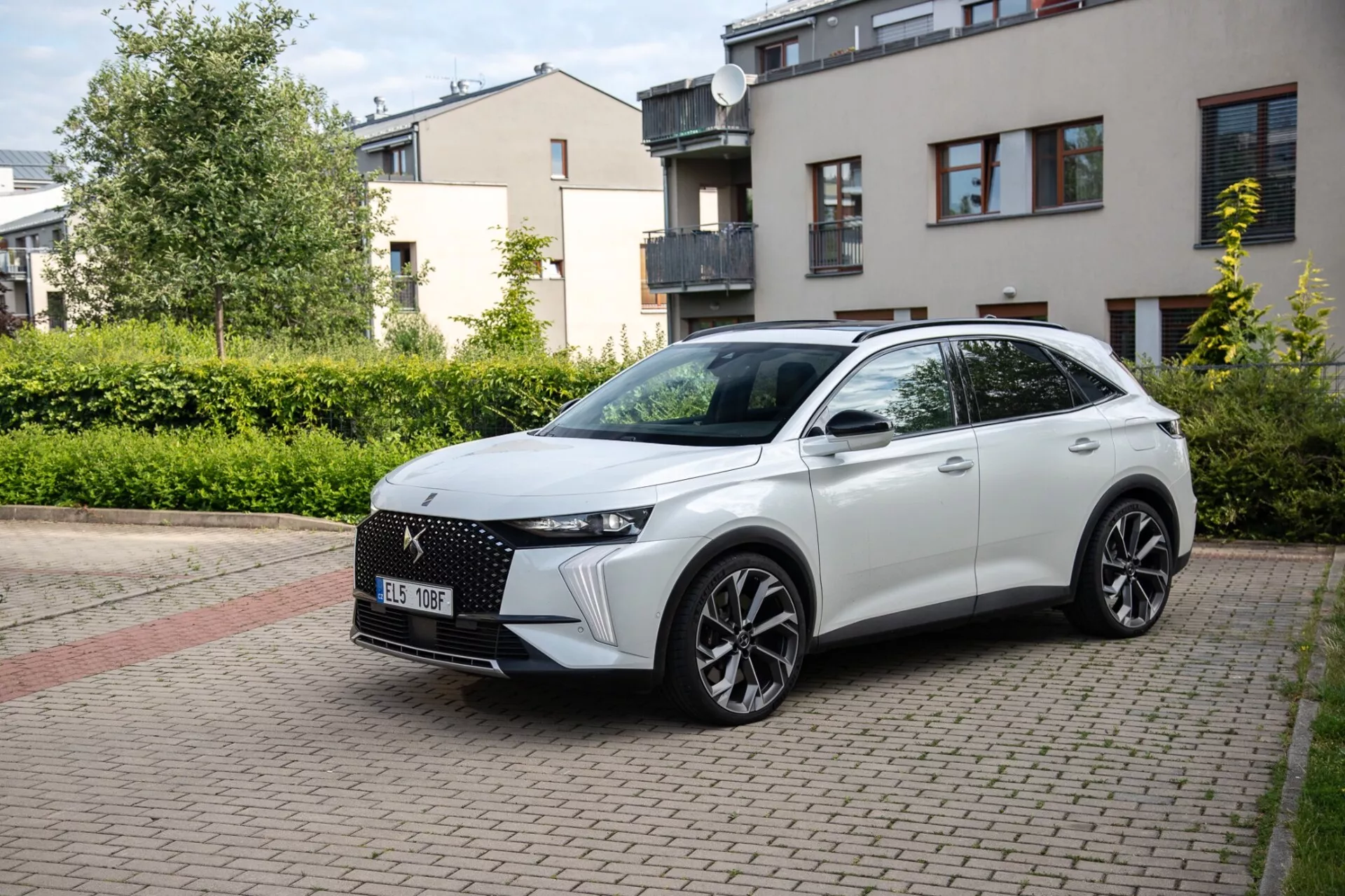 Tříčtvrteční přední pohled na kříženec DS Automobiles DS-7 2022 zaparkovaný na ulici s cihlovou dlažbou v obytné oblasti, předvádějící jeho moderní design včetně výrazné přední mřížky, elegantních LED světlometů, tvarované kapoty, velkých slitinových kol a sportovního bočního profilu s integrovanými směrovými signály na dveřích namontovaných na zrcátkách za přirozeného denního světla.