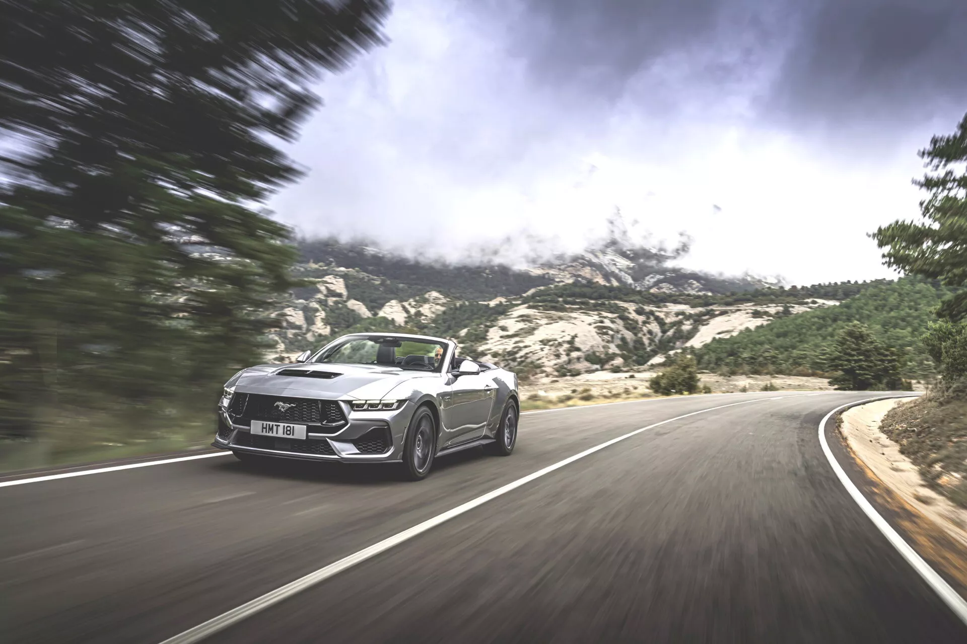 Dynamický snímek kabrioletu Ford Mustang 2024 v pohybu na klikaté horské silnici, předvádějící sportovní design a luxusní prvky na dramatickém pozadí.