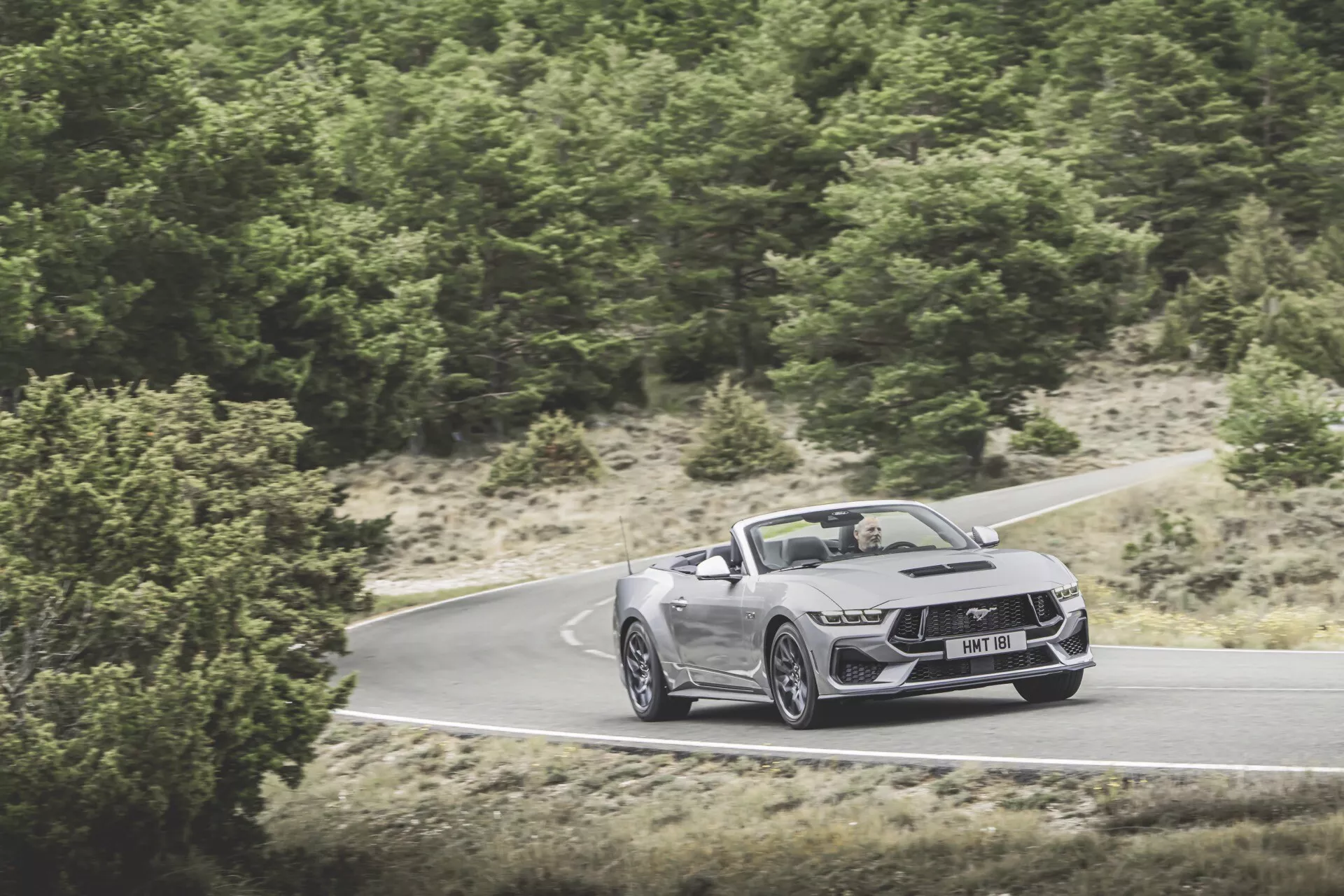 Ford Mustang Convertible 2024 v pohybu na klikaté cestě, předvádí přední část, kapotu a boční profil s aerodynamickým designem, LED světlomety a velkými slitinovými koly